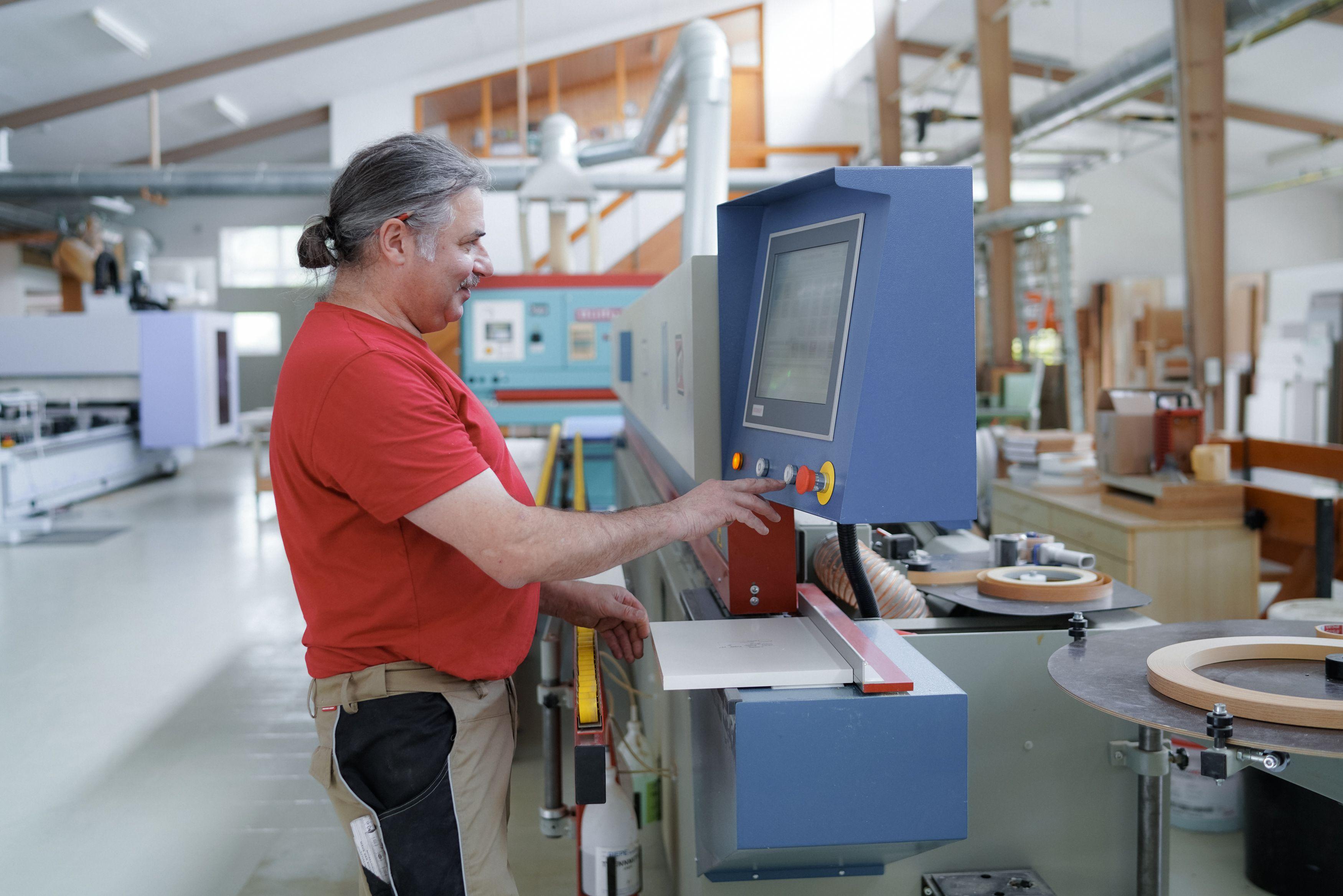 Handwerker mit grauem Zopf und rotem T-Shirt bedient eine blaue CNC-Maschine in moderner Schreinerwerkstatt mit Holzbalken-Dachkonstruktion.