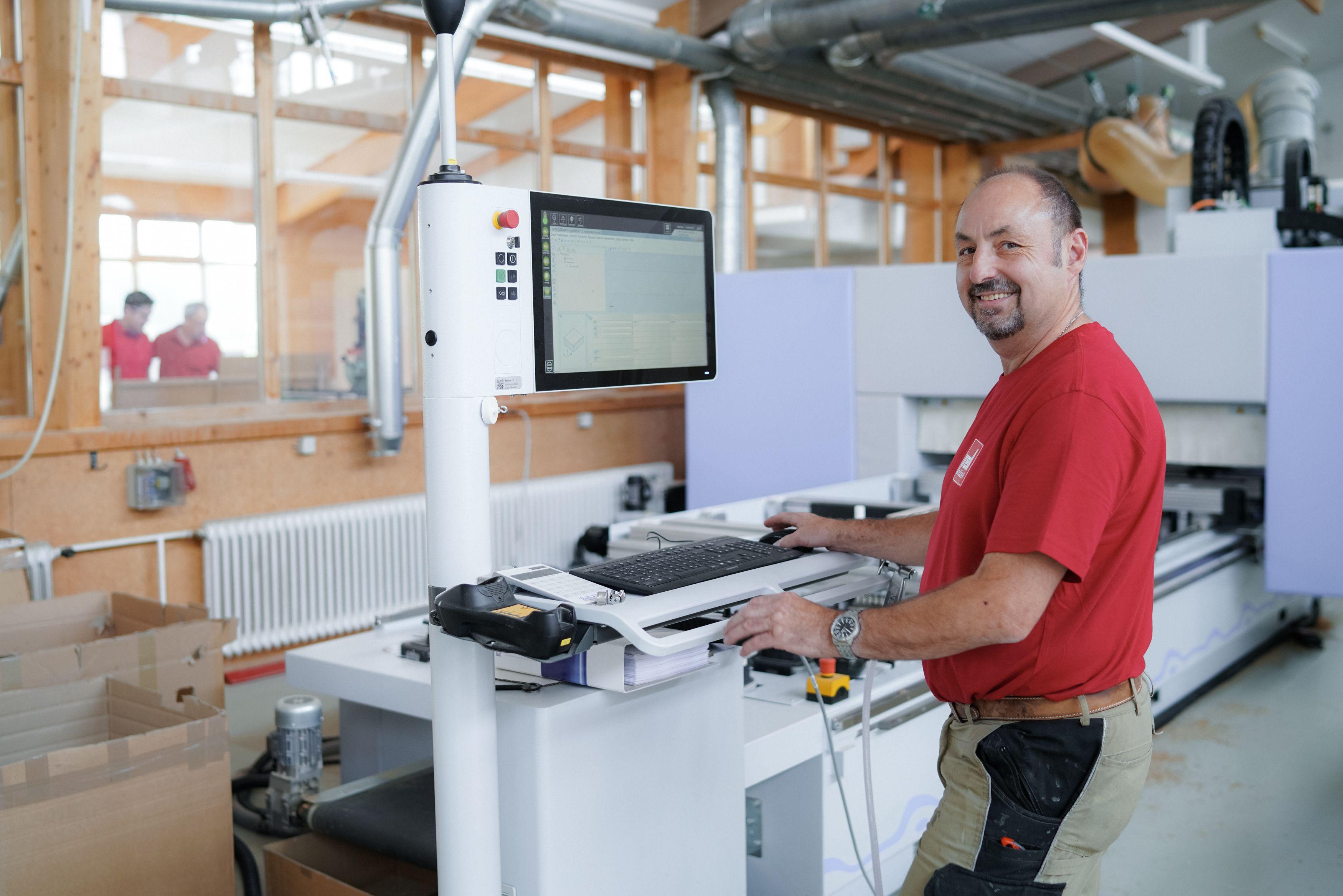 Lächelnder Arbeiter in rotem T-Shirt bedient CNC-Maschine mit Touchscreen-Monitor in moderner Holzwerkstatt mit Fachwerkkonstruktion.