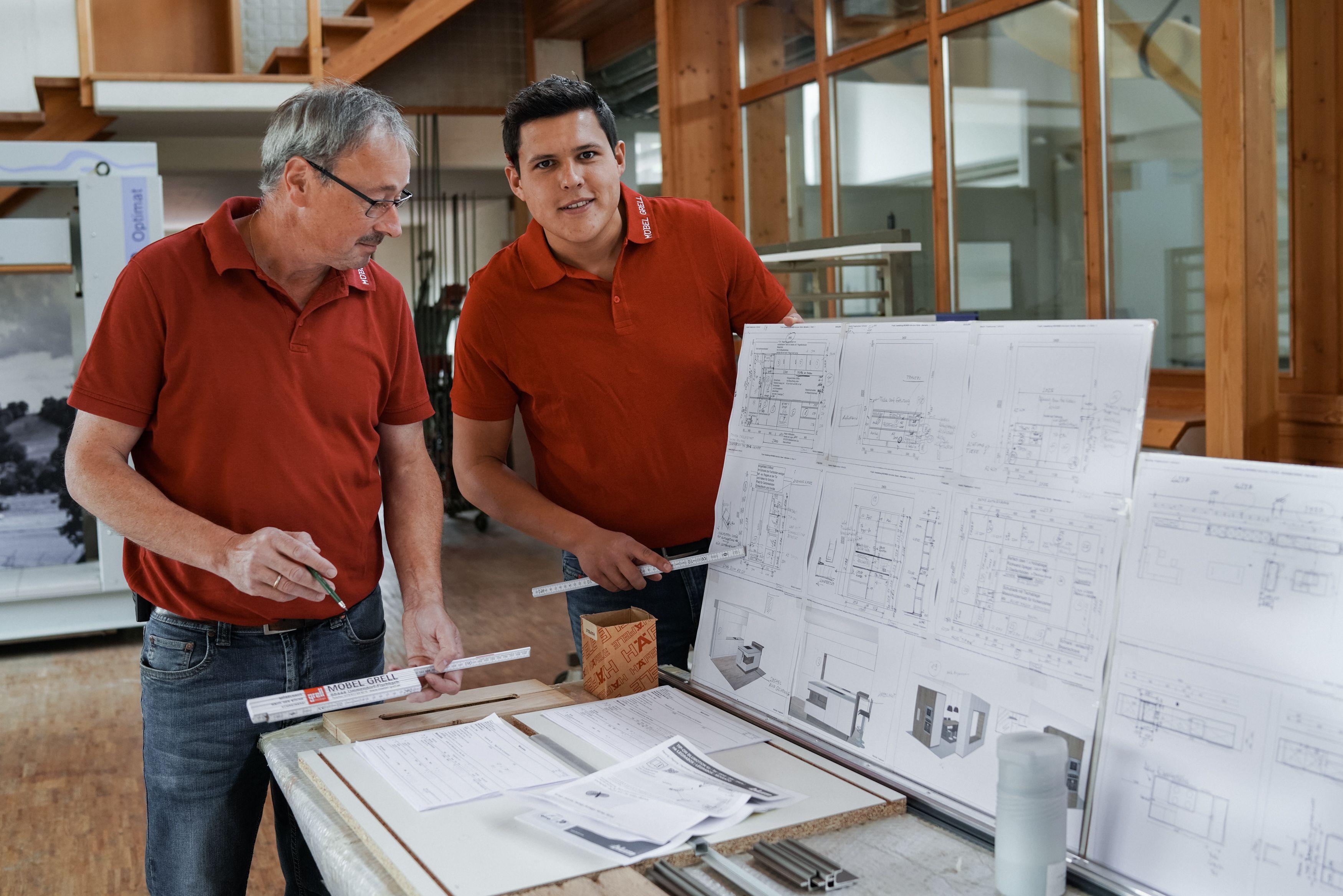 Zwei Männer in roten Poloshirts betrachten Baupläne an einem Arbeitstisch in einer Werkstatt mit Holzkonstruktion im Hintergrund.