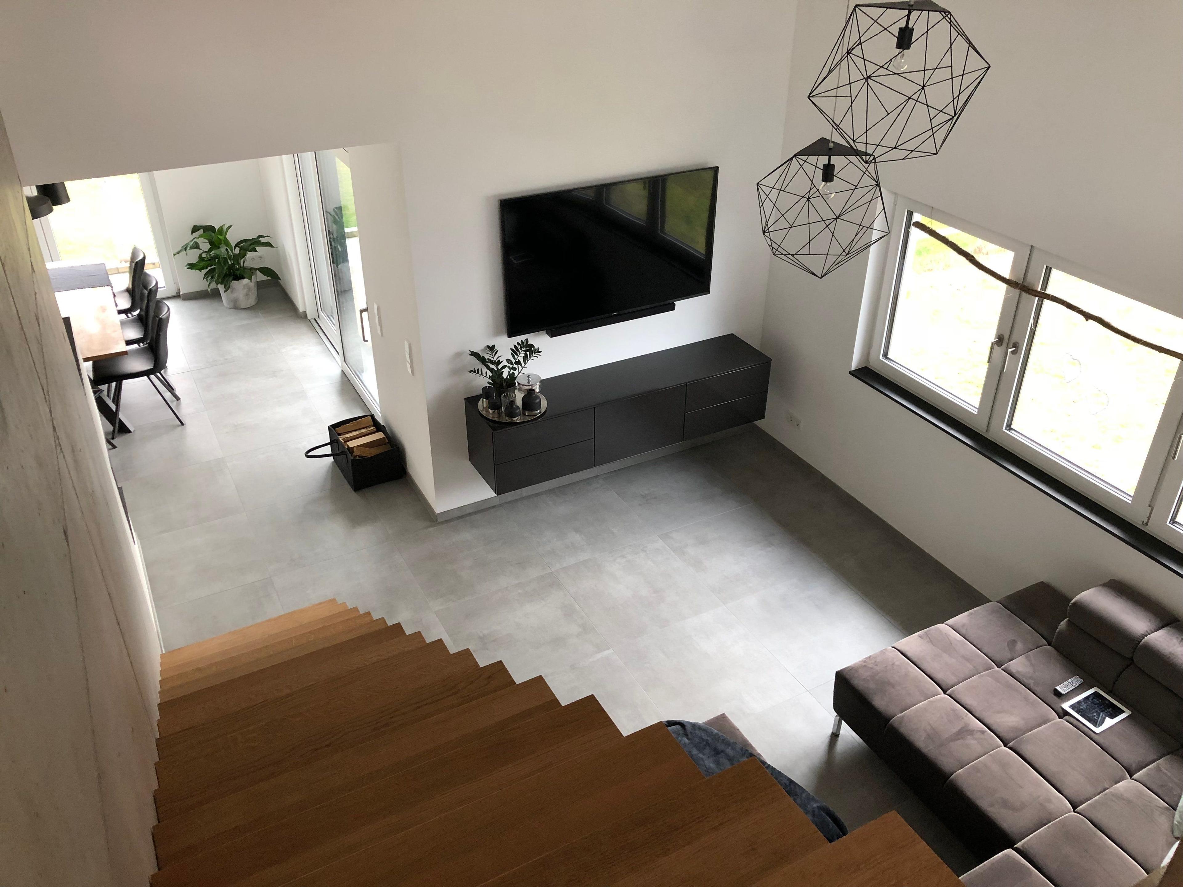 Blick von der Galerie in den Wohnbereich mit grauem Sofa, Wandfernseher, schwarzem Sideboard und geometrischen Pendelleuchten, freitragende Holztreppe sichtbar.