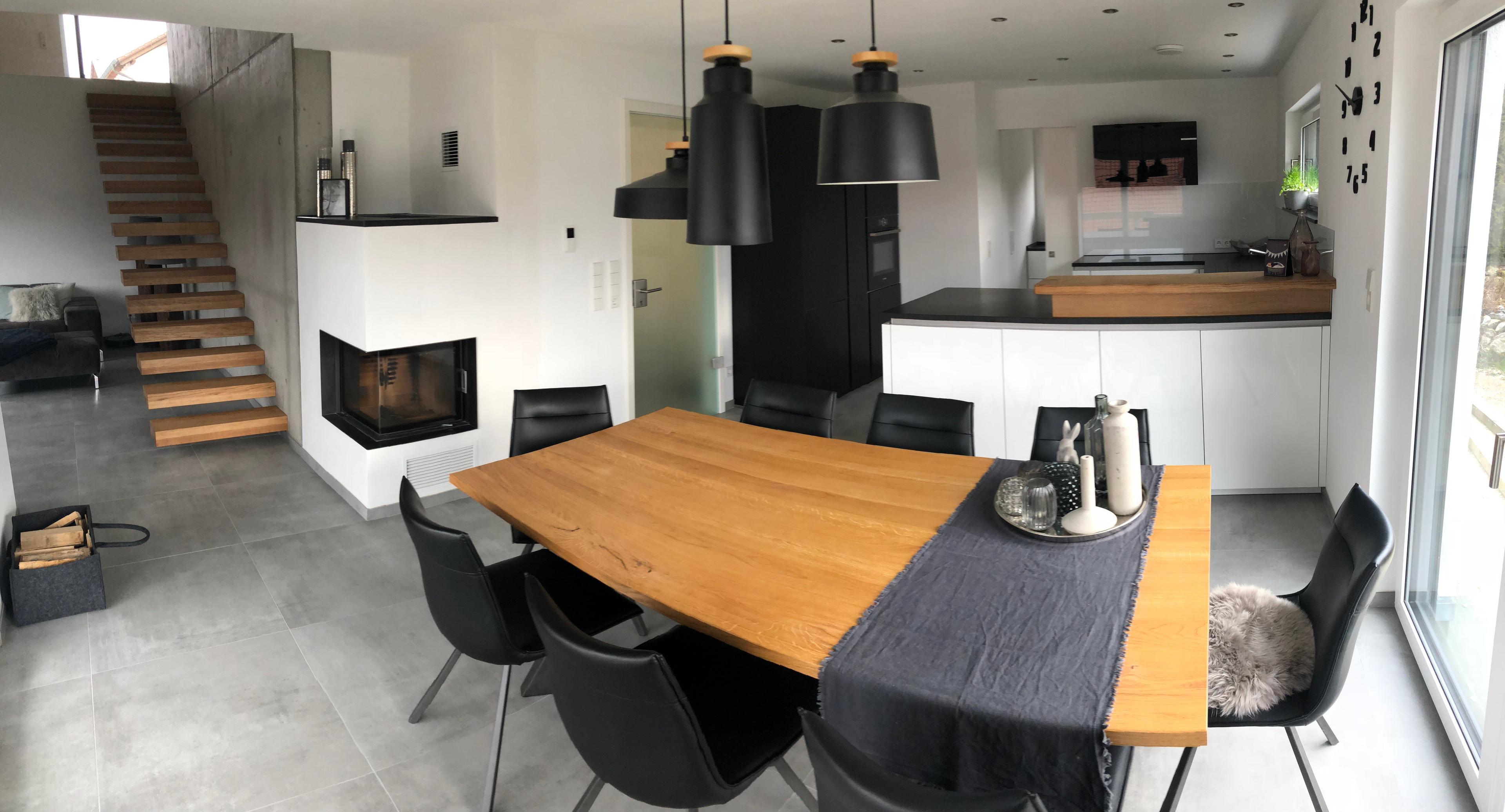 Offener Wohn-Essbereich mit modernem Kamin, Holztreppe, weißer Küche mit schwarzen Hängelampen und großem Holzesstisch mit schwarzen Stühlen.