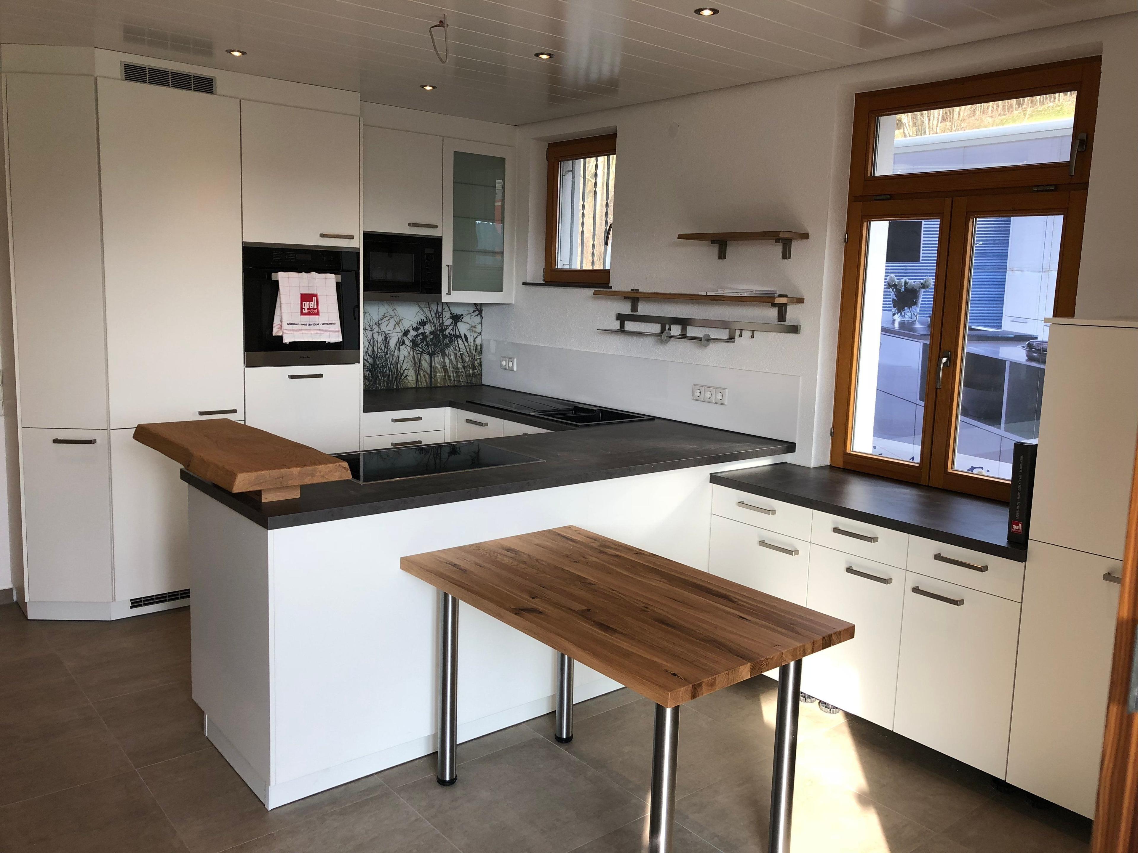 Moderne weiße Einbauküche mit schwarzer Arbeitsplatte, Kochinsel mit Holz-Schneidebrett und separatem Holztisch auf Metallbeinen, integrierte Geräte und Wandregale.