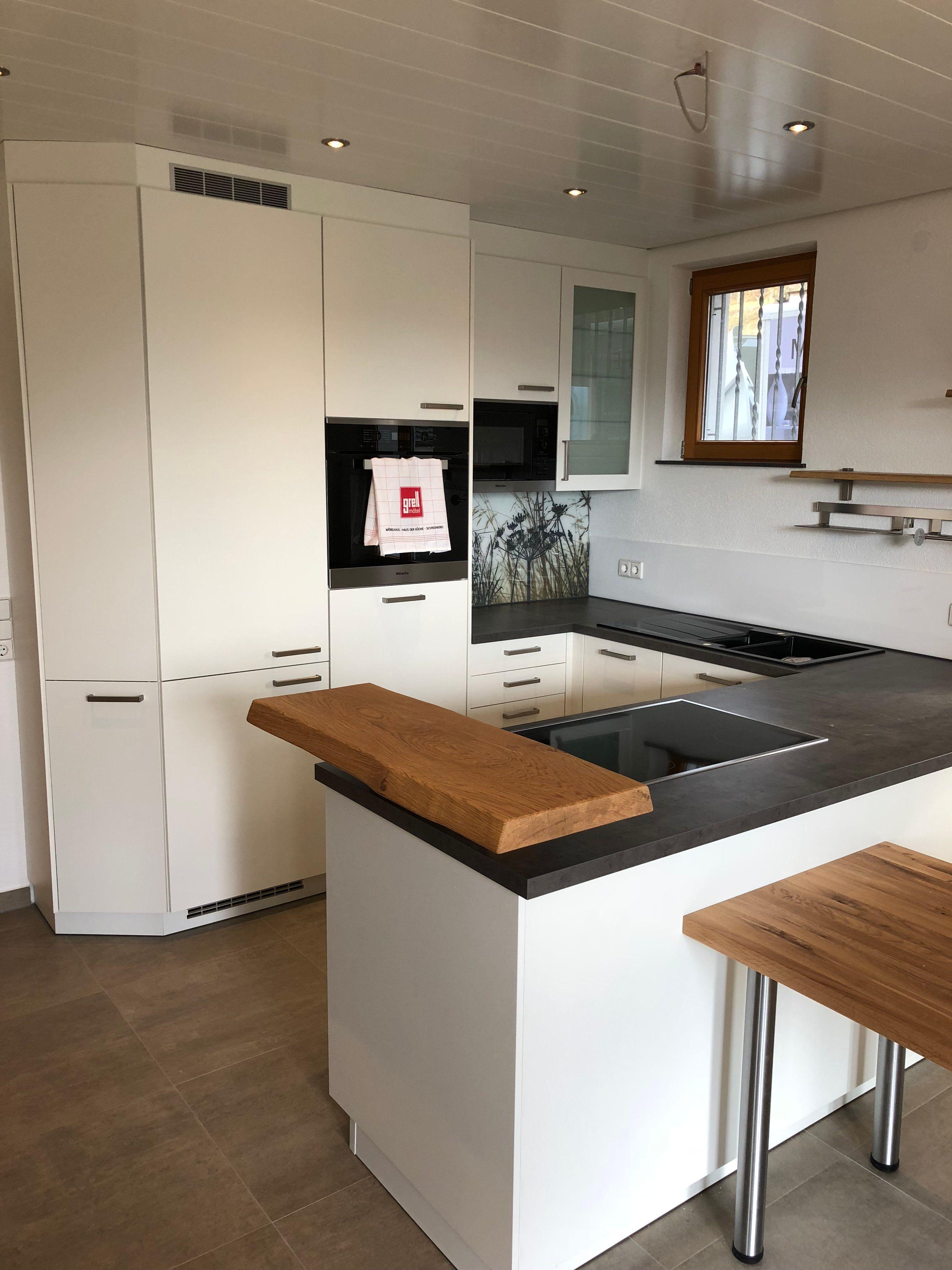 Detailansicht derselben Küche mit L-förmigem Grundriss, weißen Fronten, schwarzer Arbeitsplatte mit Induktionskochfeld und Holzelementen als Akzente