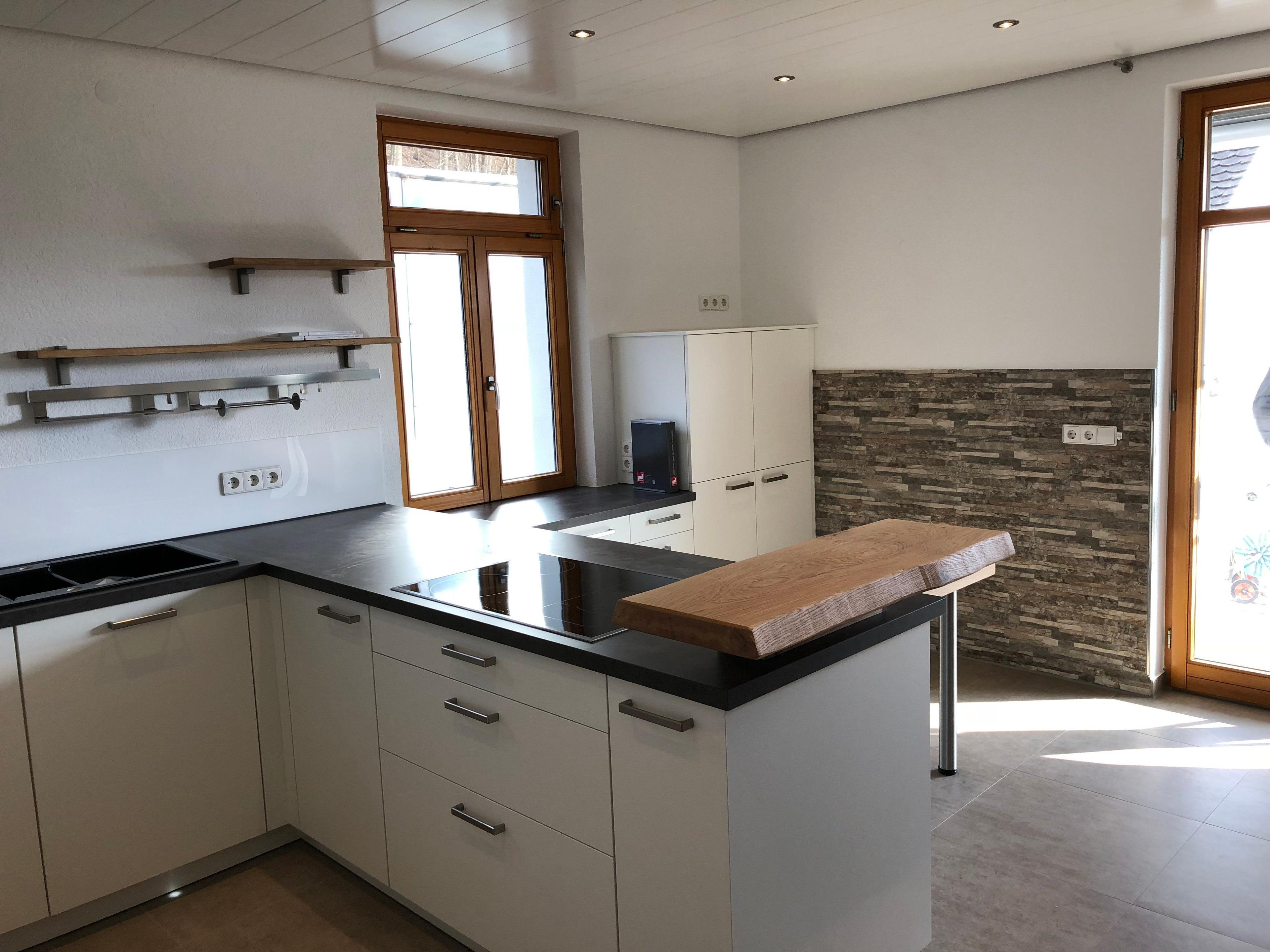 Küche mit weißen Unterschränken, schwarzer Arbeitsplatte, Natursteinwand als Rückspritzschutz und Holz-Theke an der Kücheninsel