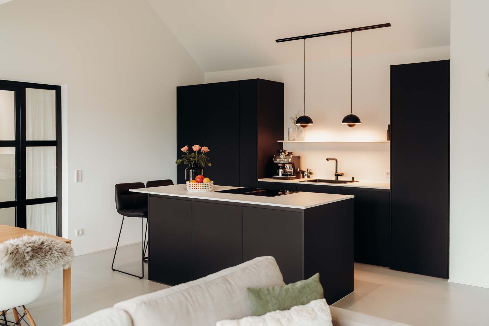Moderne offene Küche mit schwarzer Kochinsel, weißer Arbeitsplatte und zwei schwarzen Pendelleuchten, verbunden mit einem hellen Wohnbereich mit beigem Sofa.