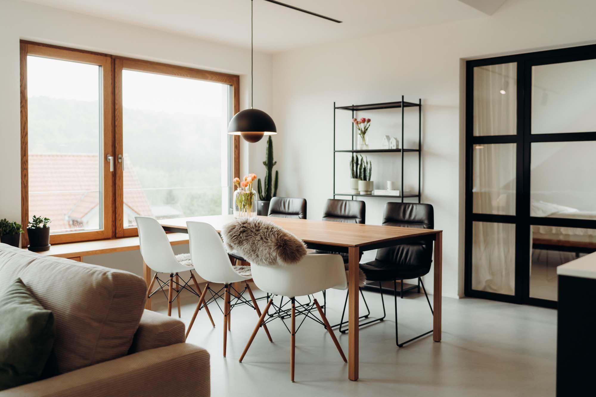 Heller Essbereich mit Holztisch, weißen und schwarzen Stühlen, schwarzer Pendelleuchte und einem schwarzen Metallregal mit Pflanzen und Geschirr im Hintergrund.