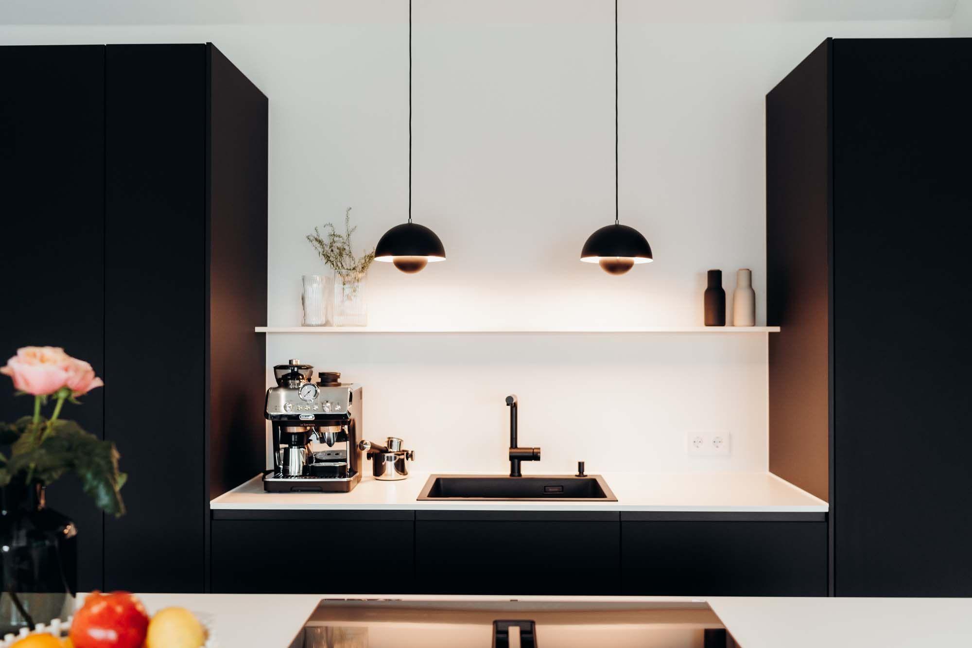 Küchenarbeitsbereich mit schwarzen Oberschränken, weißer Arbeitsplatte, Espressomaschine, zwei schwarzen Pendelleuchten und minimalistischer weißer Rückwand.