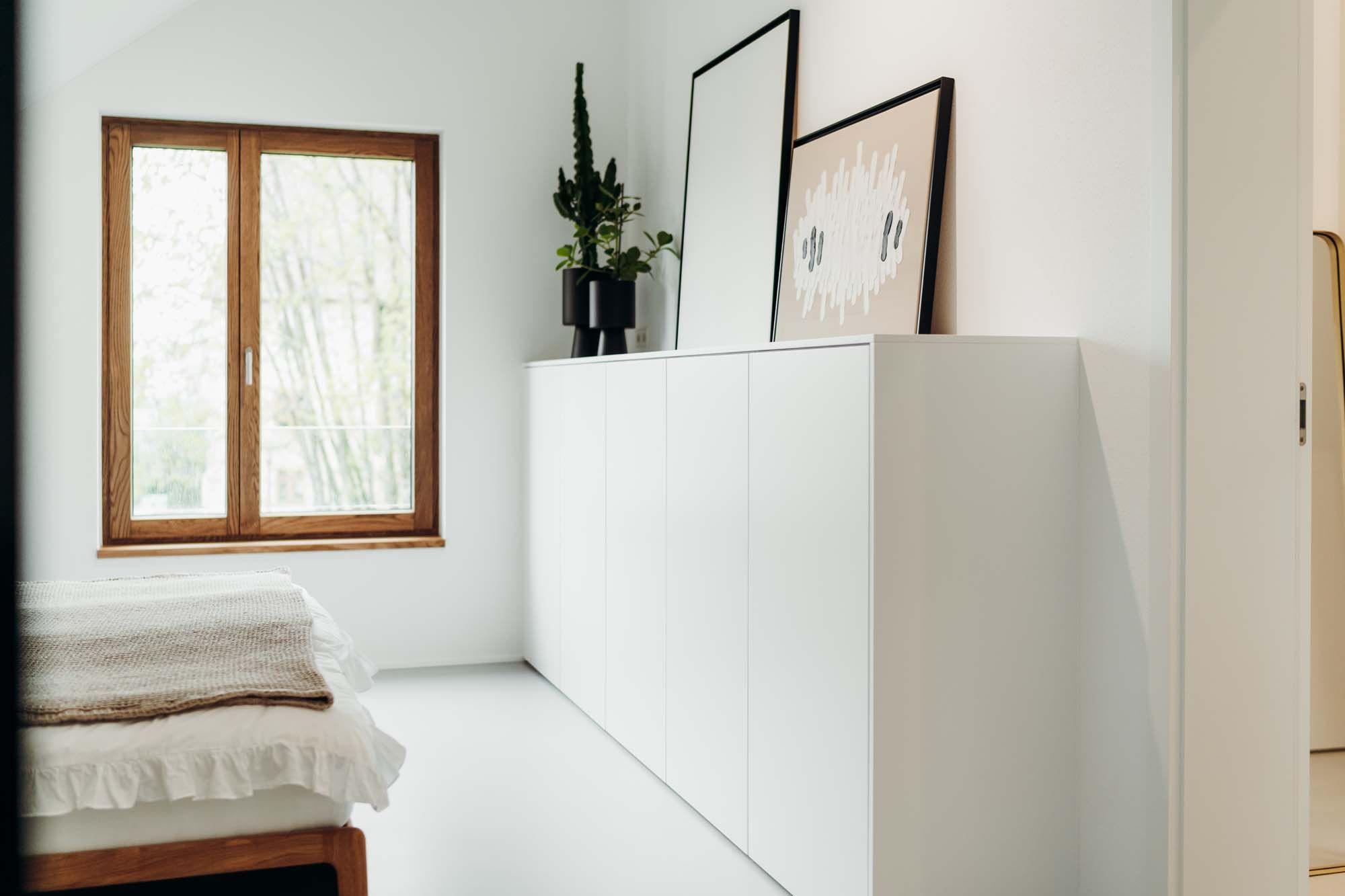 Weißes Sideboard in einem hellen Raum mit zwei gerahmten Bildern darauf, einer schwarzen Pflanze und Blick in einen angrenzenden Raum.