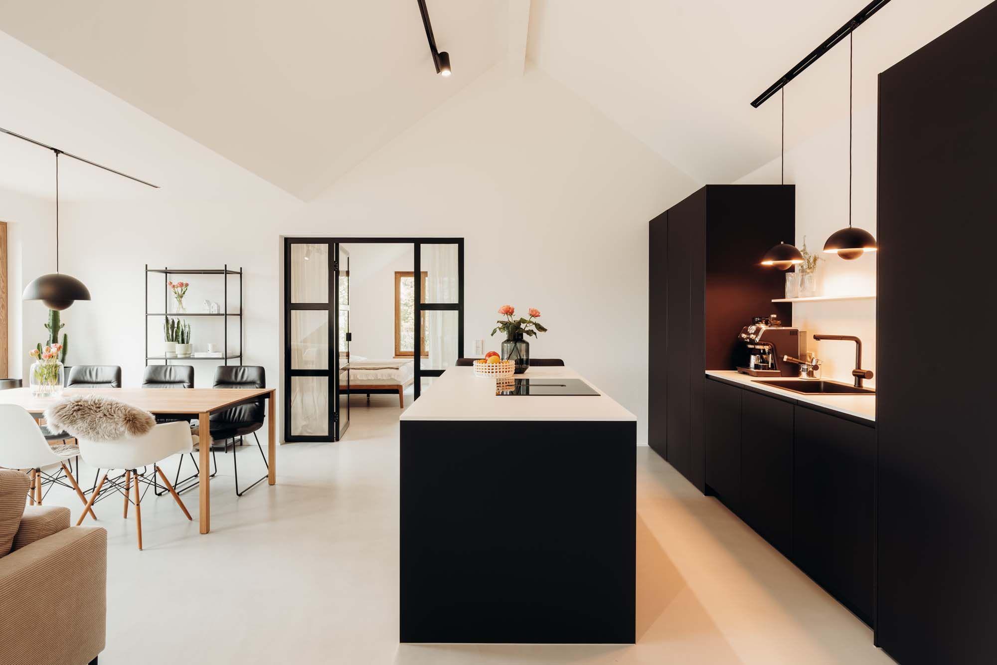 Weitläufiger offener Wohnbereich mit schwarzer Küche, Kochinsel, Essbereich mit Holztisch, weißen Designerstühlen und großen Glasfronten.