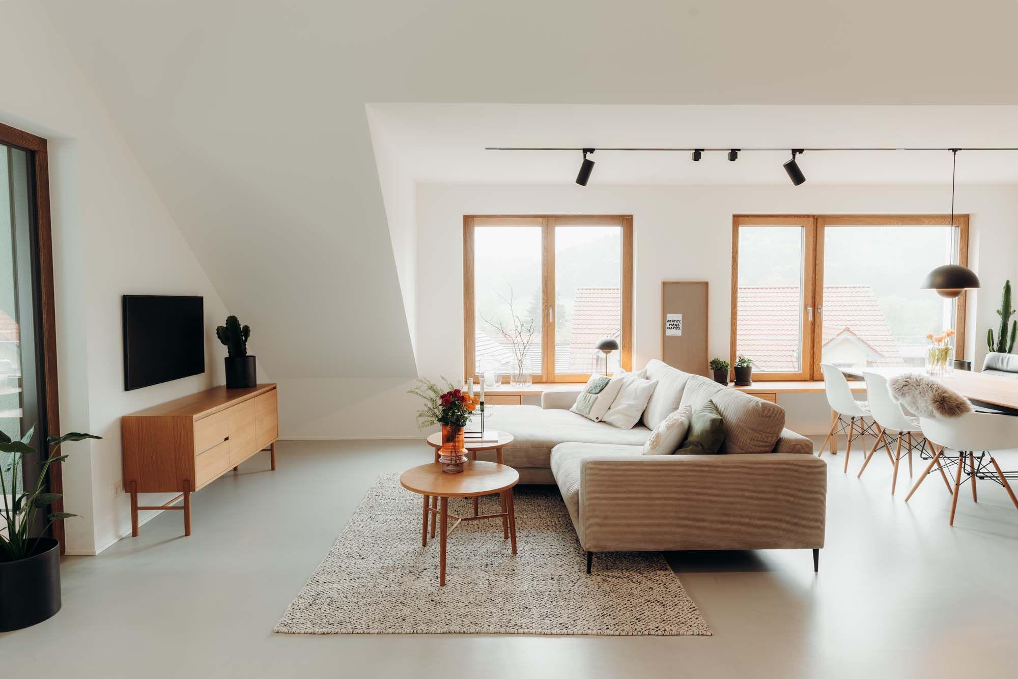Helles Dachgeschoss-Wohnzimmer mit beigem Sofa, Holzcouchtisch, TV-Möbel, schwarzem Fernseher und großen Holzfenstern mit Blick auf Dächer.