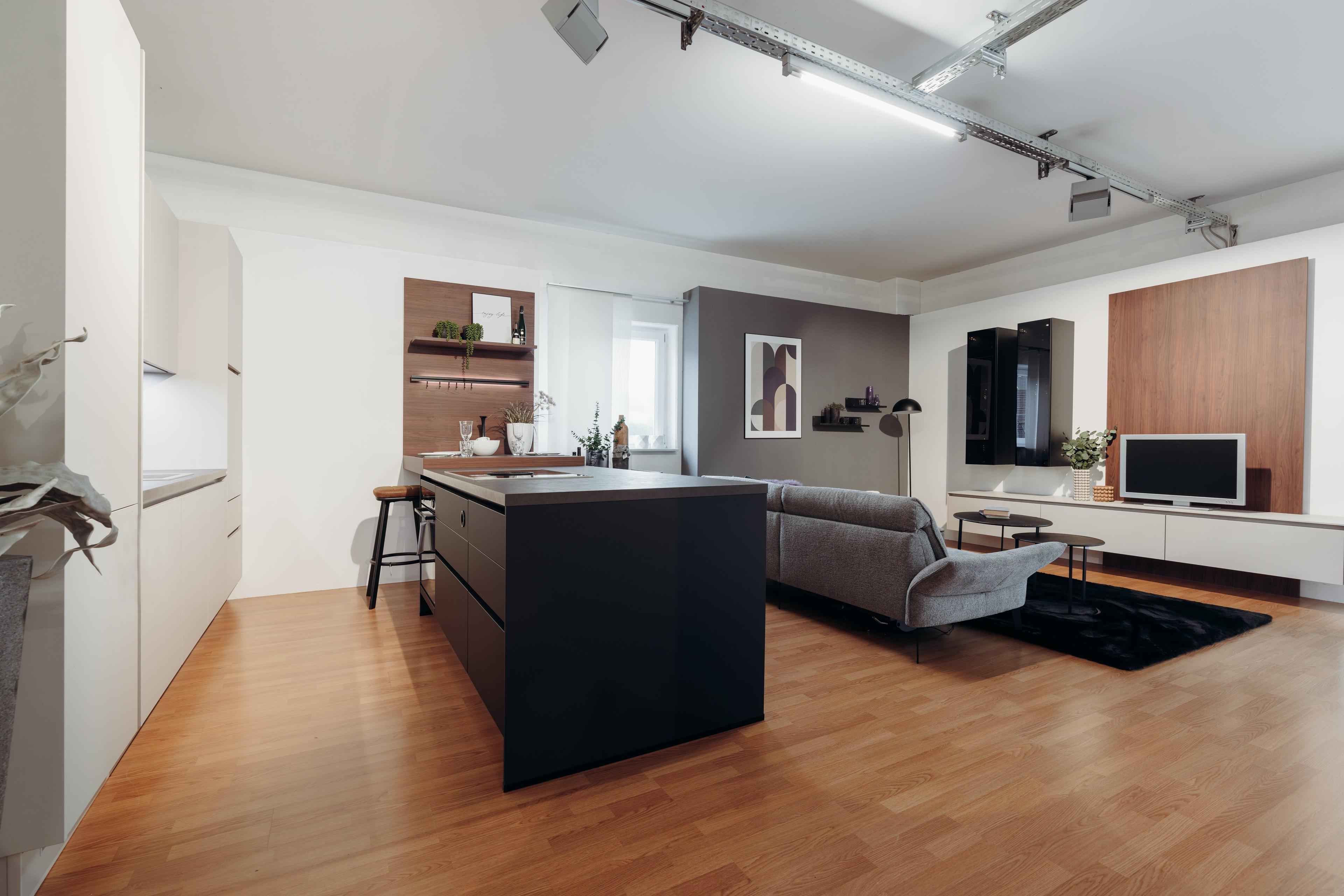 Offener Wohn-Küchenbereich mit schwarzer Kücheninsel, weißen Fronten und grauem Sofa vor TV-Wand aus Holz und weißen Elementen auf Parkettboden.