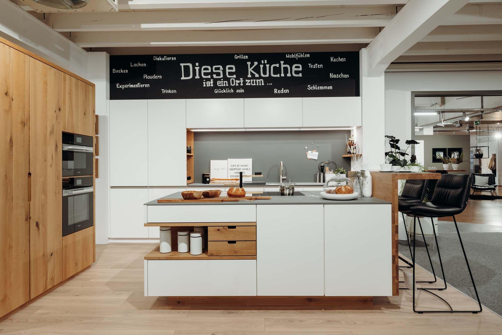 Moderne Küche mit weißen Fronten und Holzdetails, ergänzt durch eine Wand mit inspirierendem Text und stilvollen Barhockern. Der Küchenbereich zeigt eine offene, einladende Atmosphäre.
