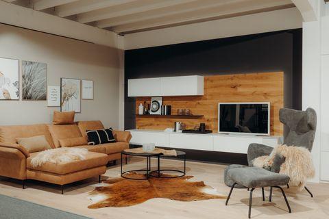 Moderne Wohnzimmer-Ausstellung mit cognacfarbenem Ledersofa, TV-Wand aus Holz und weißen Elementen, grauem Sessel und Kuhfell-Teppich.