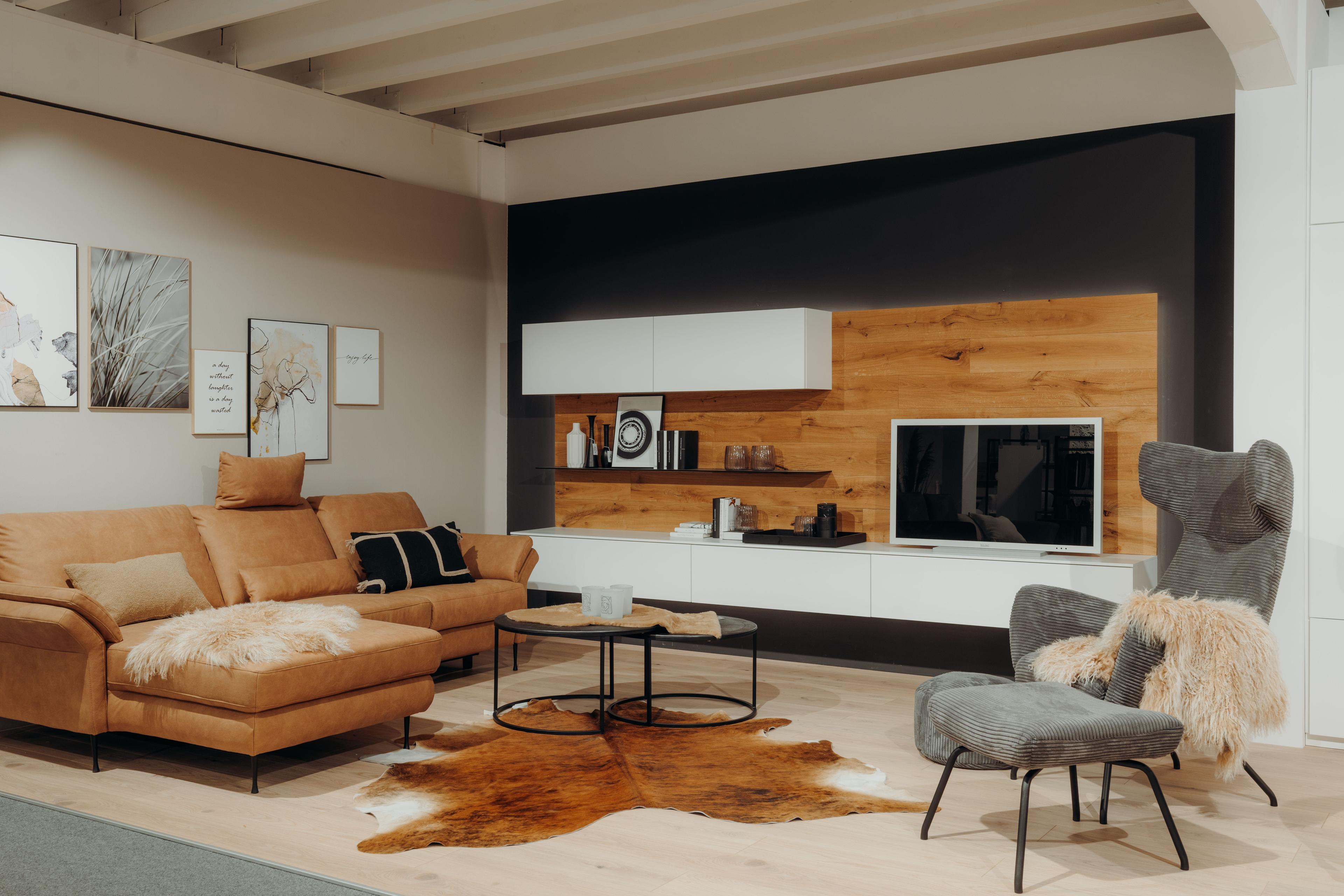 Moderne Wohnzimmer-Ausstellung mit cognacfarbenem Ledersofa, TV-Wand aus Holz und weißen Elementen, grauem Sessel und Kuhfell-Teppich.