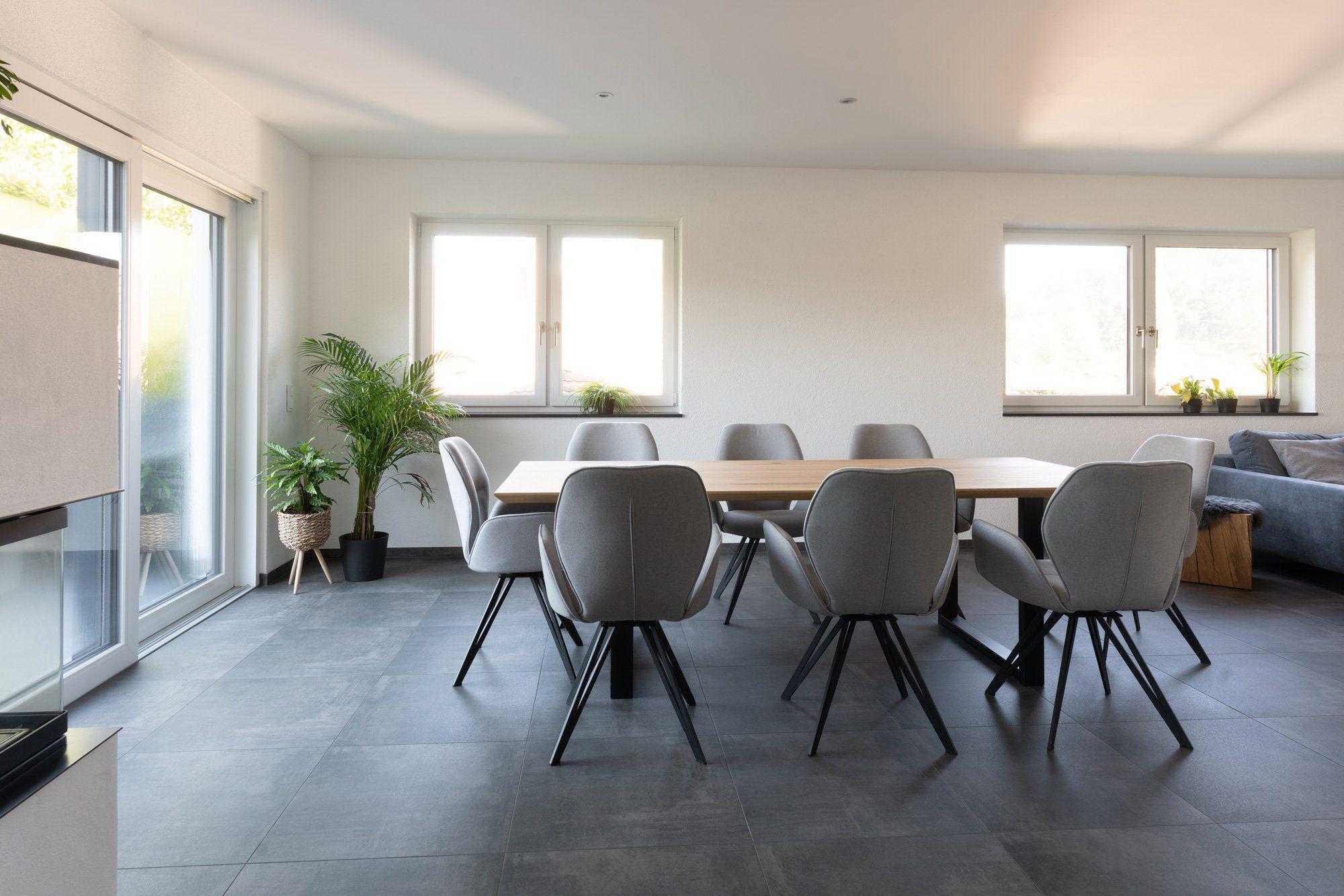 Essbereich mit Holztisch, acht grauen Stühlen, Zimmerpflanzen und großen Fenstern in hellem Wohnraum.