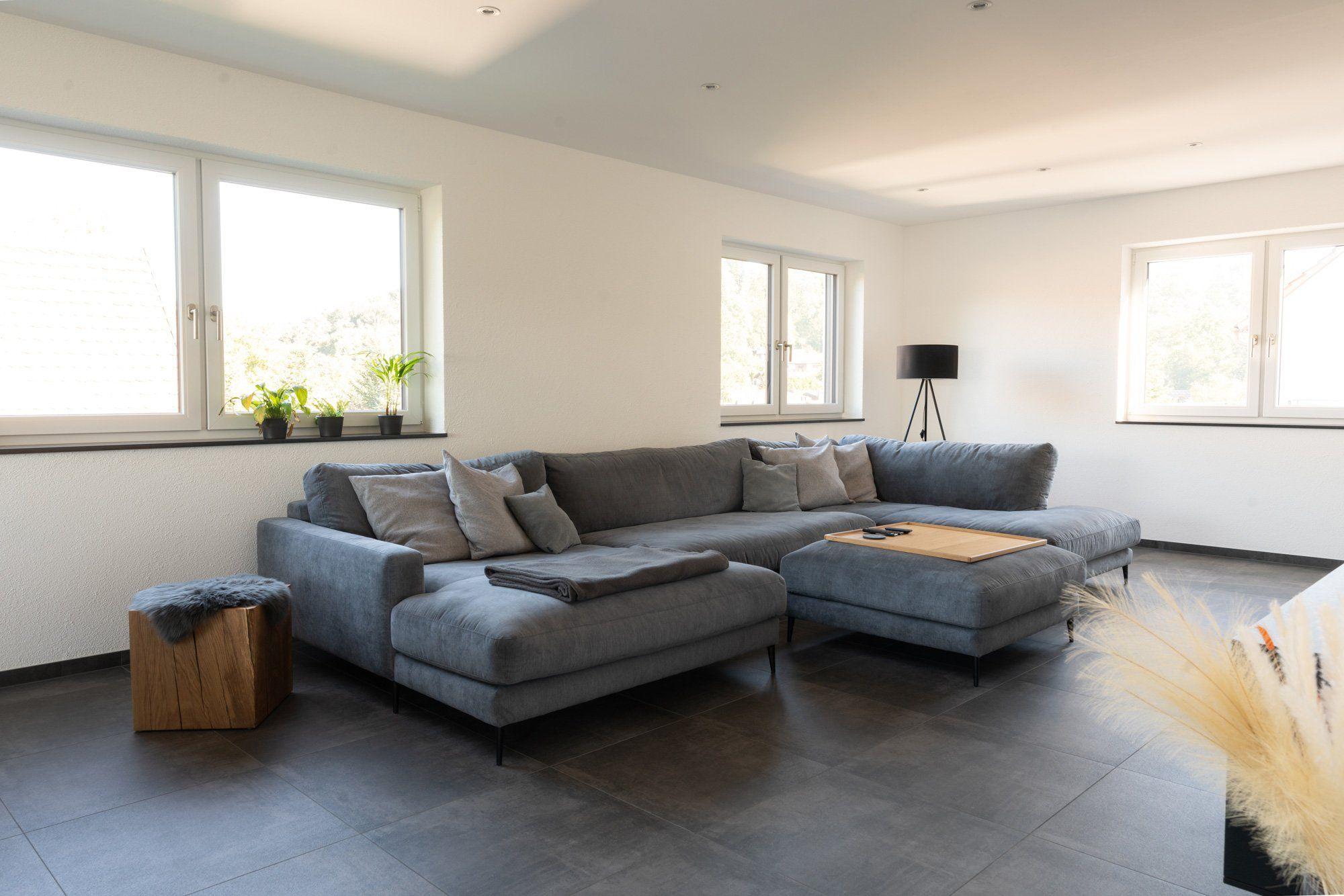 Helles Wohnzimmer mit grauer Eckcouch, Holzhocker, Stehlampe mit schwarzem Schirm und großen Fenstern mit Zimmerpflanzen.