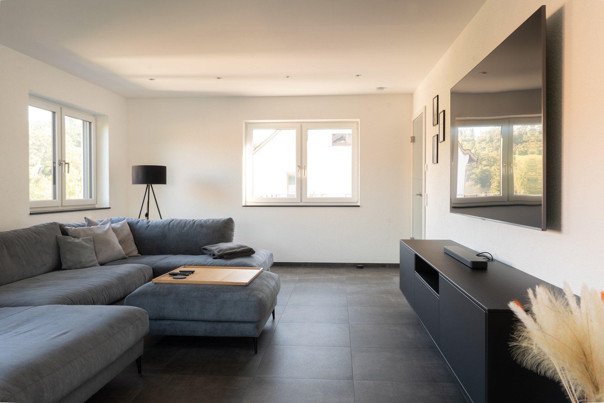 Wohnbereich mit grauem Ecksofa, schwarzem TV-Lowboard, Wandfernseher und Dreibein-Stehleuchte vor weißen Wänden.