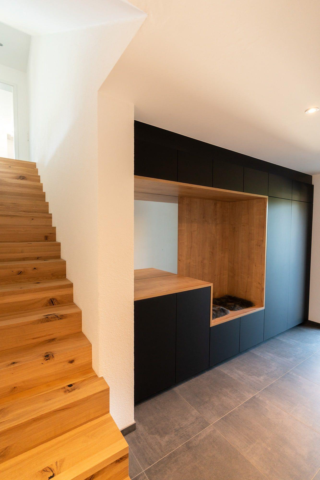 Treppenaufgang aus Eichenholz neben Einbauschrank mit Holznische, schwarzen Fronten und grauer Fliesenboden im Eingangsbereich.