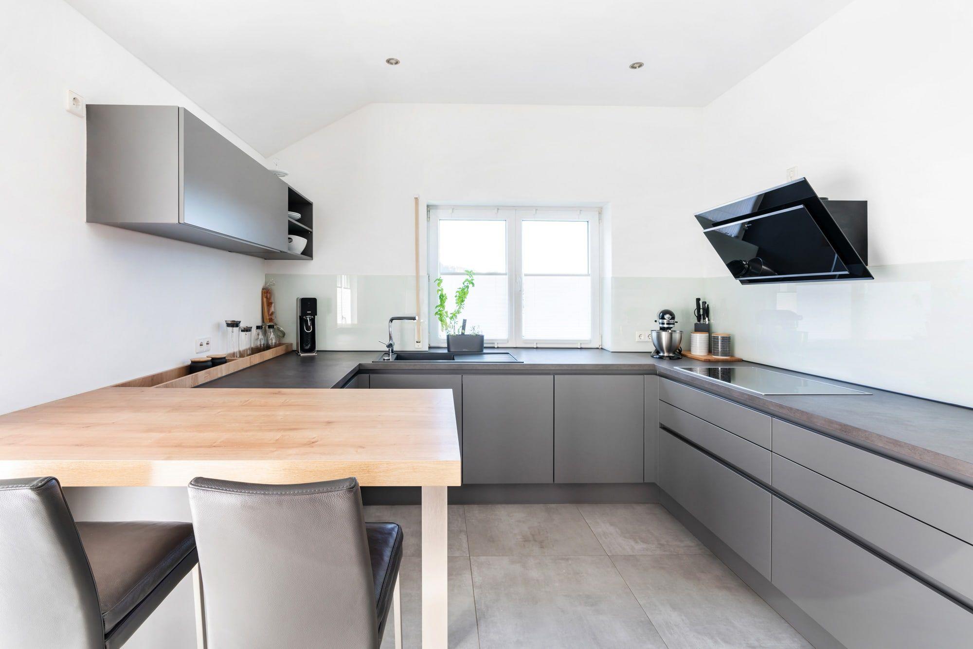 Moderne U-förmige Küche in Grau mit schwarzer Arbeitsplatte, Holz-Esstheke, schwarzer Dunstabzugshaube und beigen Barhockern.