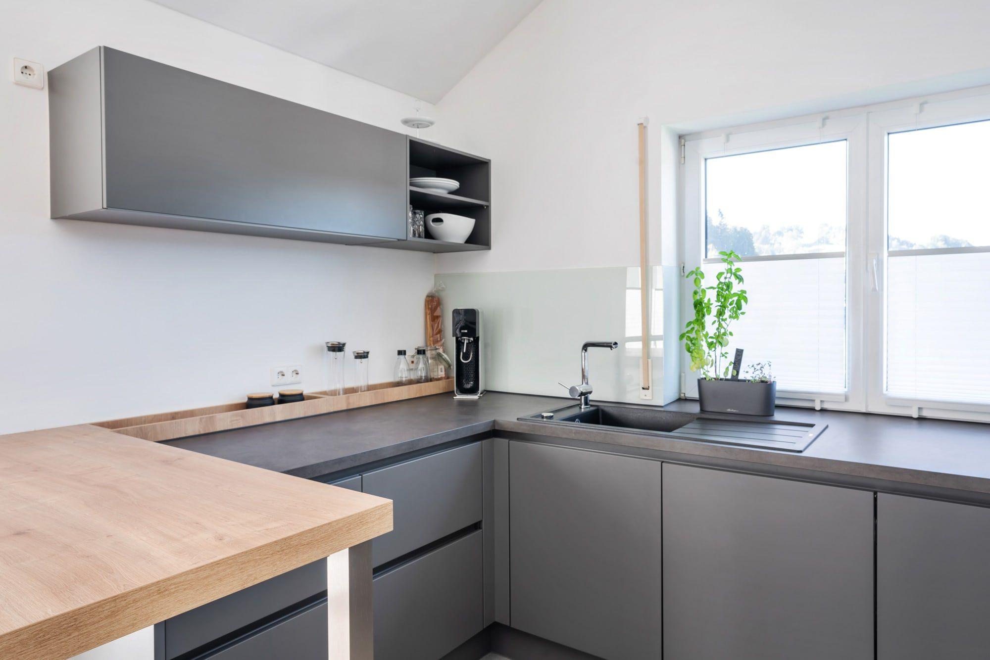 Küchenecke mit grauer Front, schwarzer Arbeitsplatte, Holzleiste als Wandabschluss und Eckfenster mit Pflanze