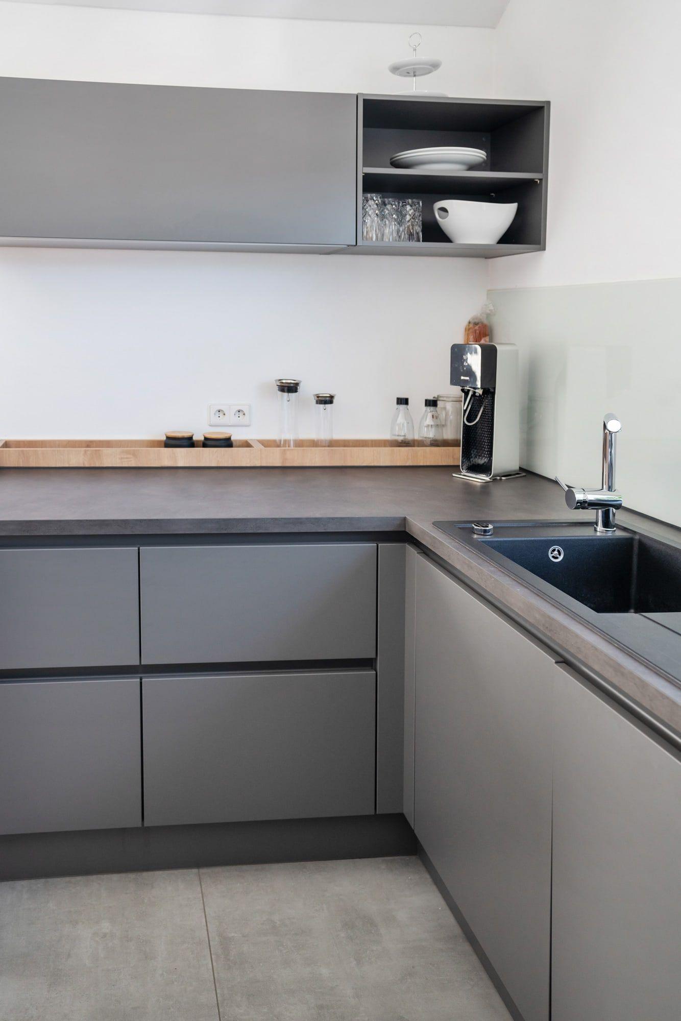 Küchendetail mit grauen Unterschränken, offenen Hängeschränken, Holzregal, schwarzer Spüle und weißer Glasrückwand.