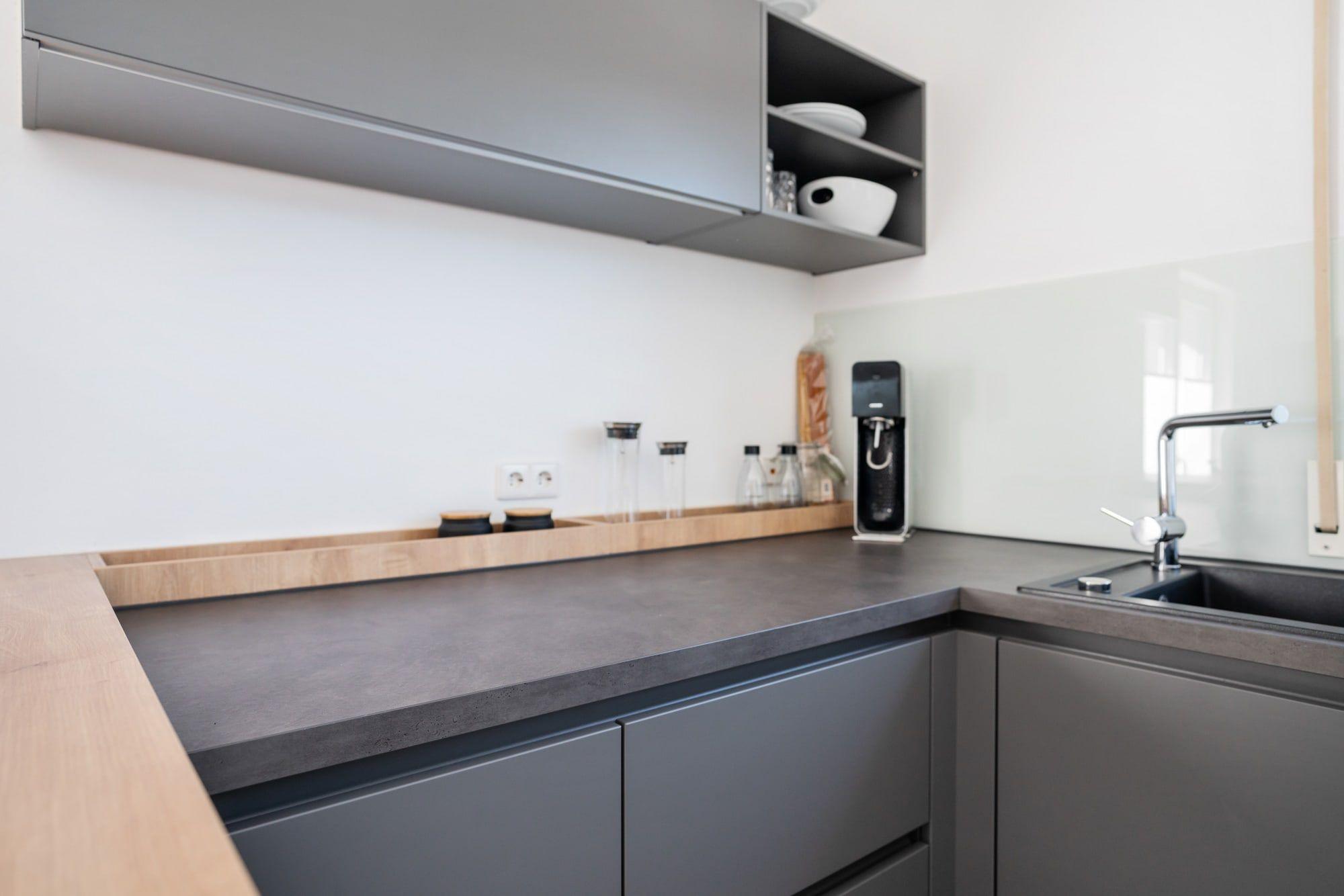 Küchenarbeitsplatz mit grauer Front, schwarzer Arbeitsplatte, Holzleiste als Wandabschluss und modernen Elektrogeräten.