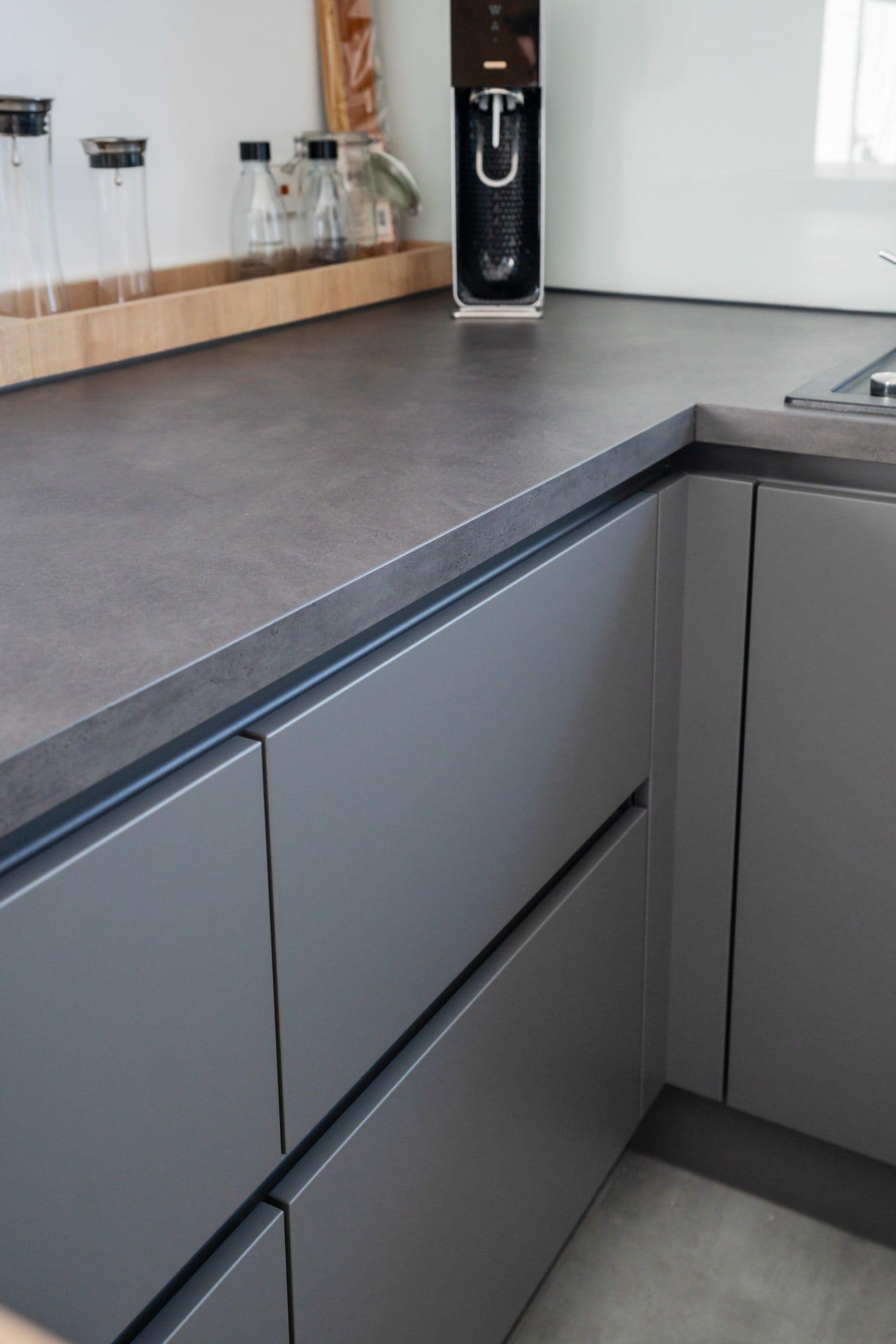 Detailansicht der Küchenarbeitsplatte mit schwarzer Oberfläche, grauen grifflosen Fronten, Holzleiste als Wandabschluss und schwarzer Kaffeemaschine.