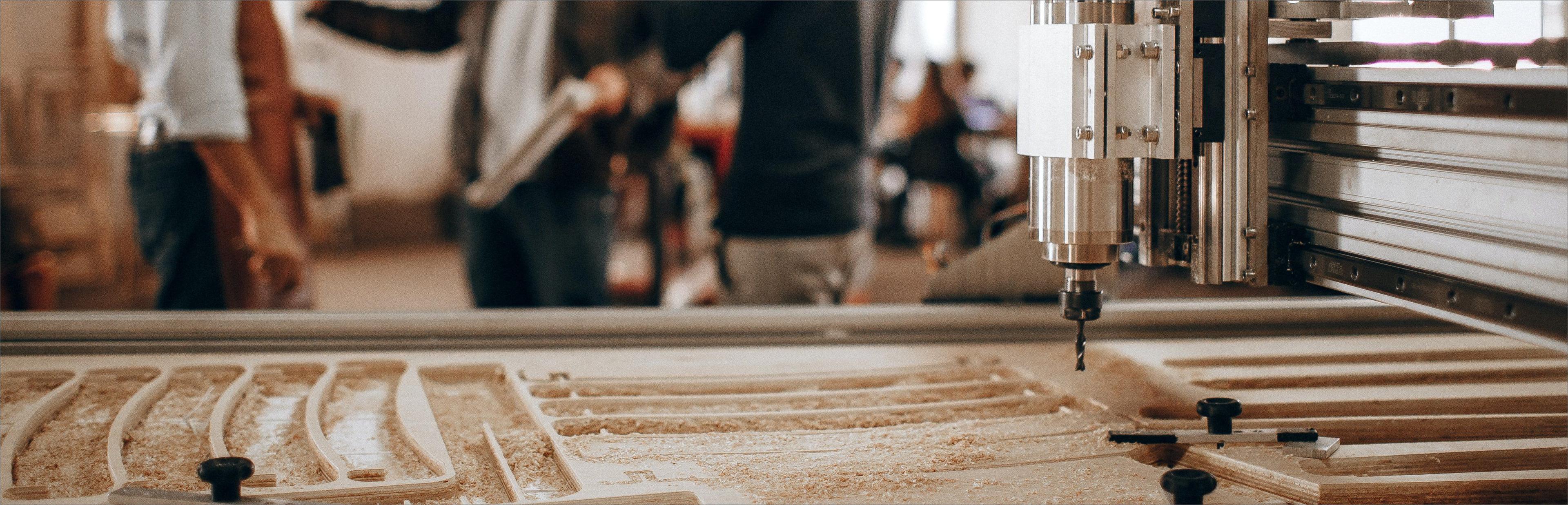 CNC-Fräsmaschine bearbeitet Holzwerkstück und produziert Sägespäne, im Hintergrund unscharf sichtbare Arbeiter in einer Werkstatt.
