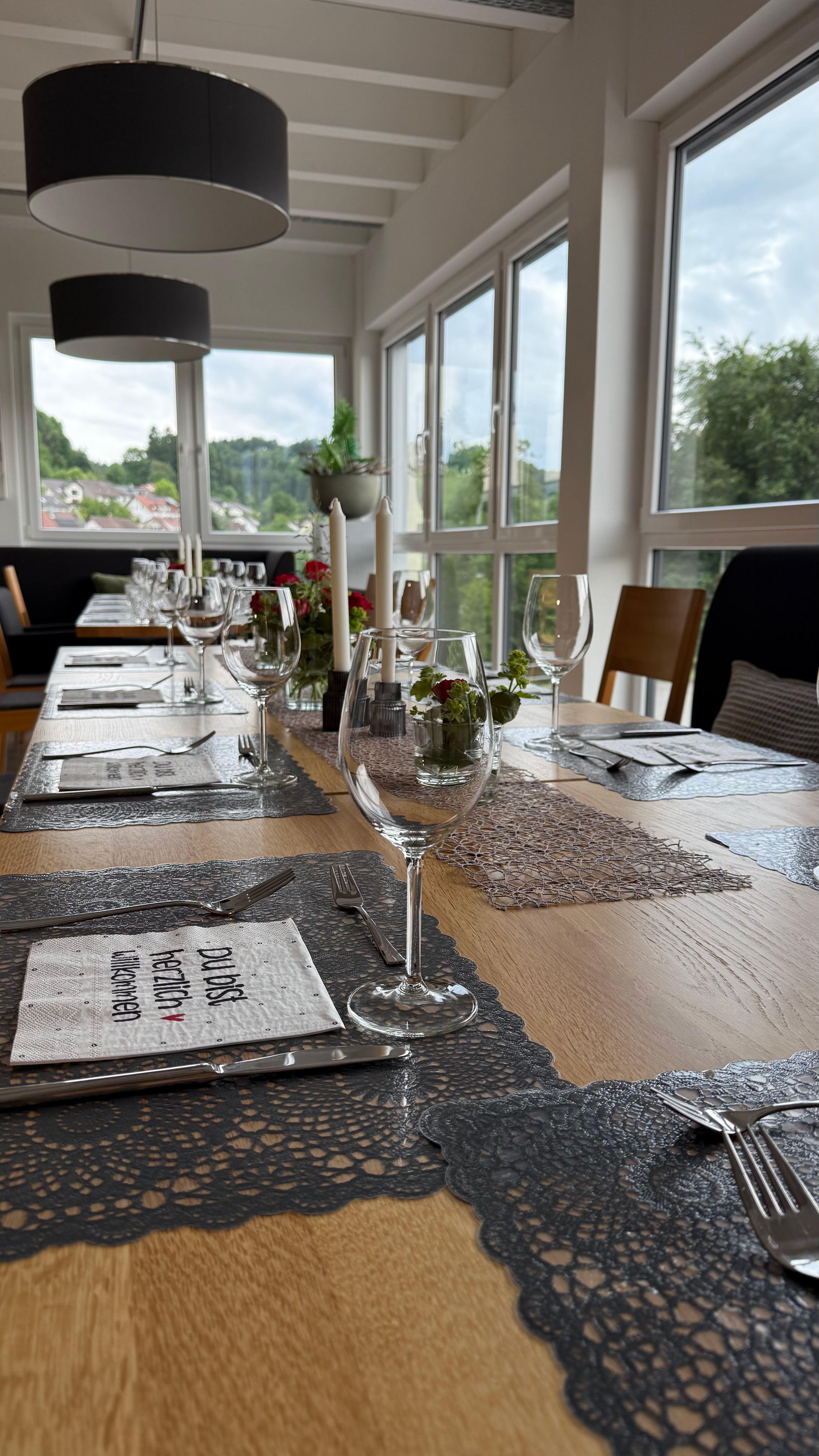 Elegant gedeckter Holztisch mit Weingläsern, schwarzen Platzsets, weißen Kerzen und großen Fenstern mit Ausblick in lichtdurchfluteten Raum.