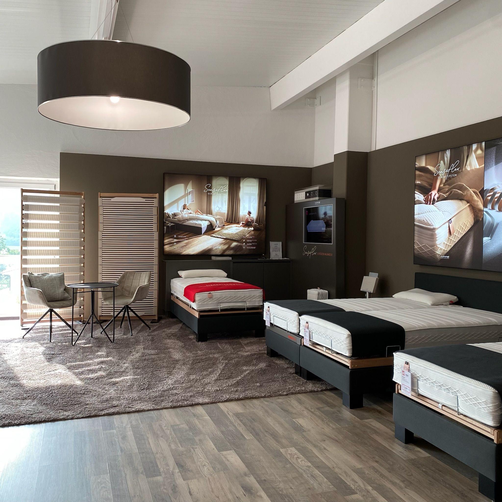 Moderner Matratzenshowroom mit mehreren ausgestellten Betten auf schwarzen Gestellen, großen Werbeplakaten an den Wänden und einer eleganten schwarzen Pendelleuchte unter sichtbaren Dachbalken.
