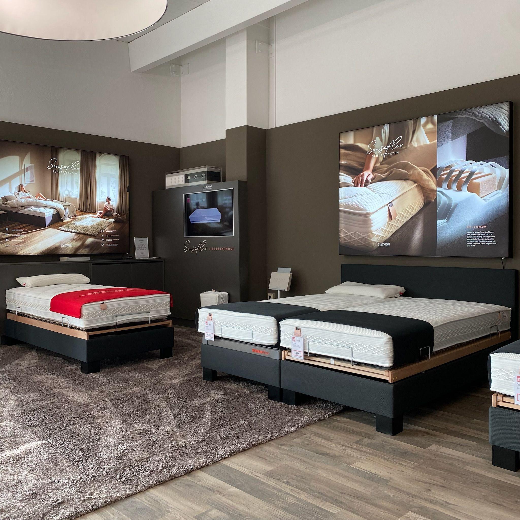 Nahaufnahme eines Bettenfachgeschäfts mit verschiedenen Matratzen auf schwarzen Plattformen, großformatigen Werbeplakaten der Marke Swissflex an der Wand und einem Bildschirm zur Produktpräsentation.