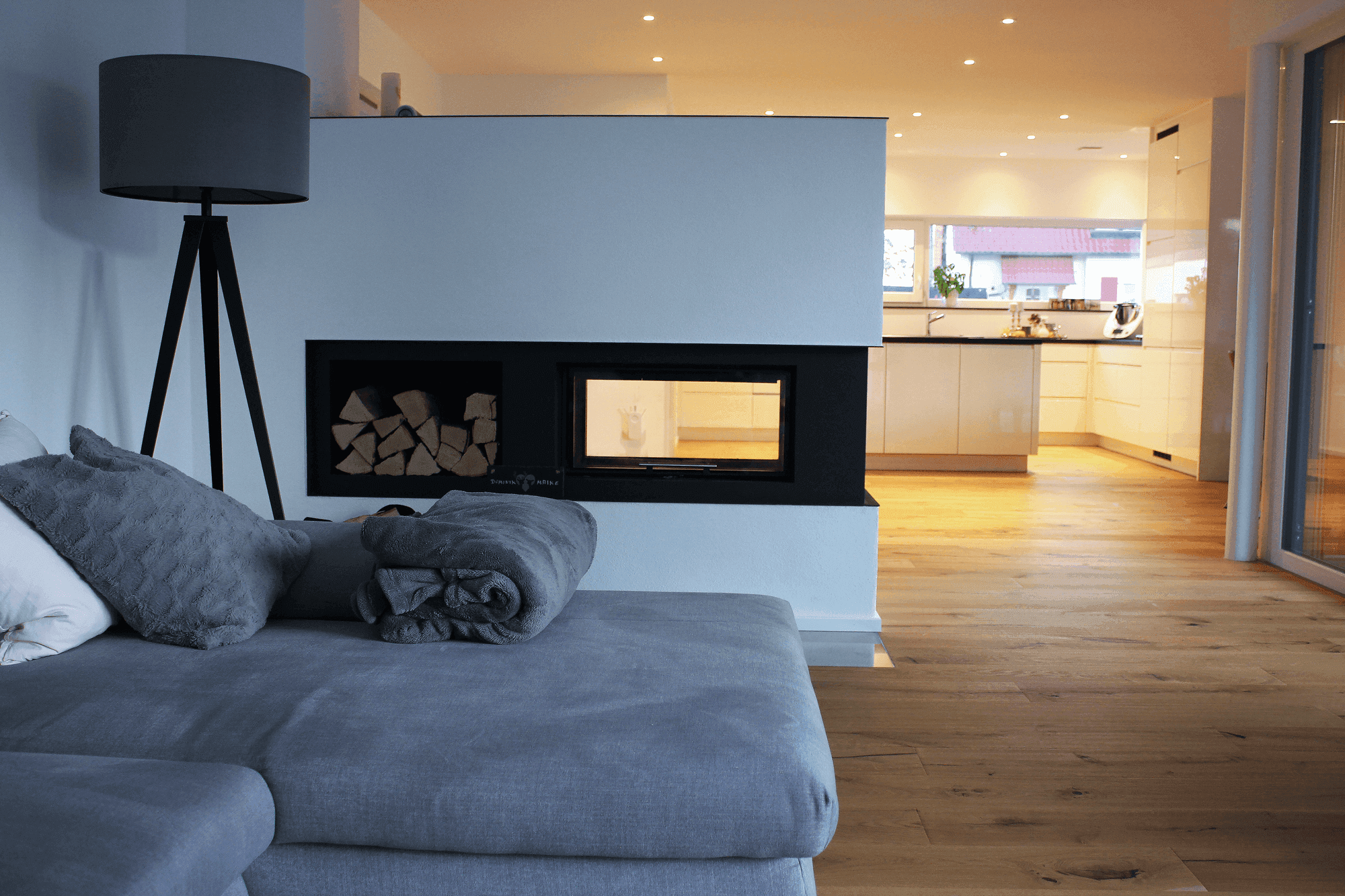 Wohnbereich mit grauem Sofa, moderner Stehlampe, Durchsichtkamin und Blick in die offene Küche.