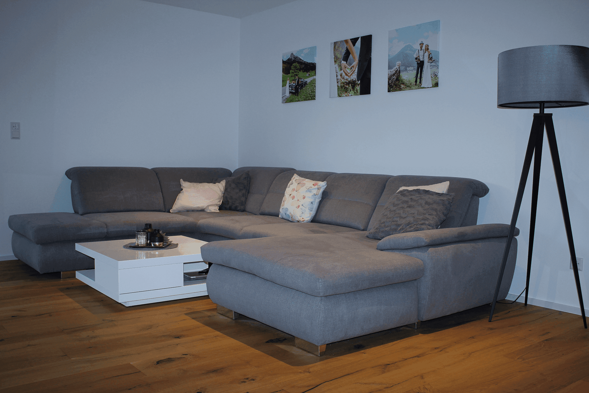 Gemütliches Wohnzimmer mit großer grauer Eckcouch, weißem Couchtisch, Stehlampe und Berglandschaft-Bildern an der Wand