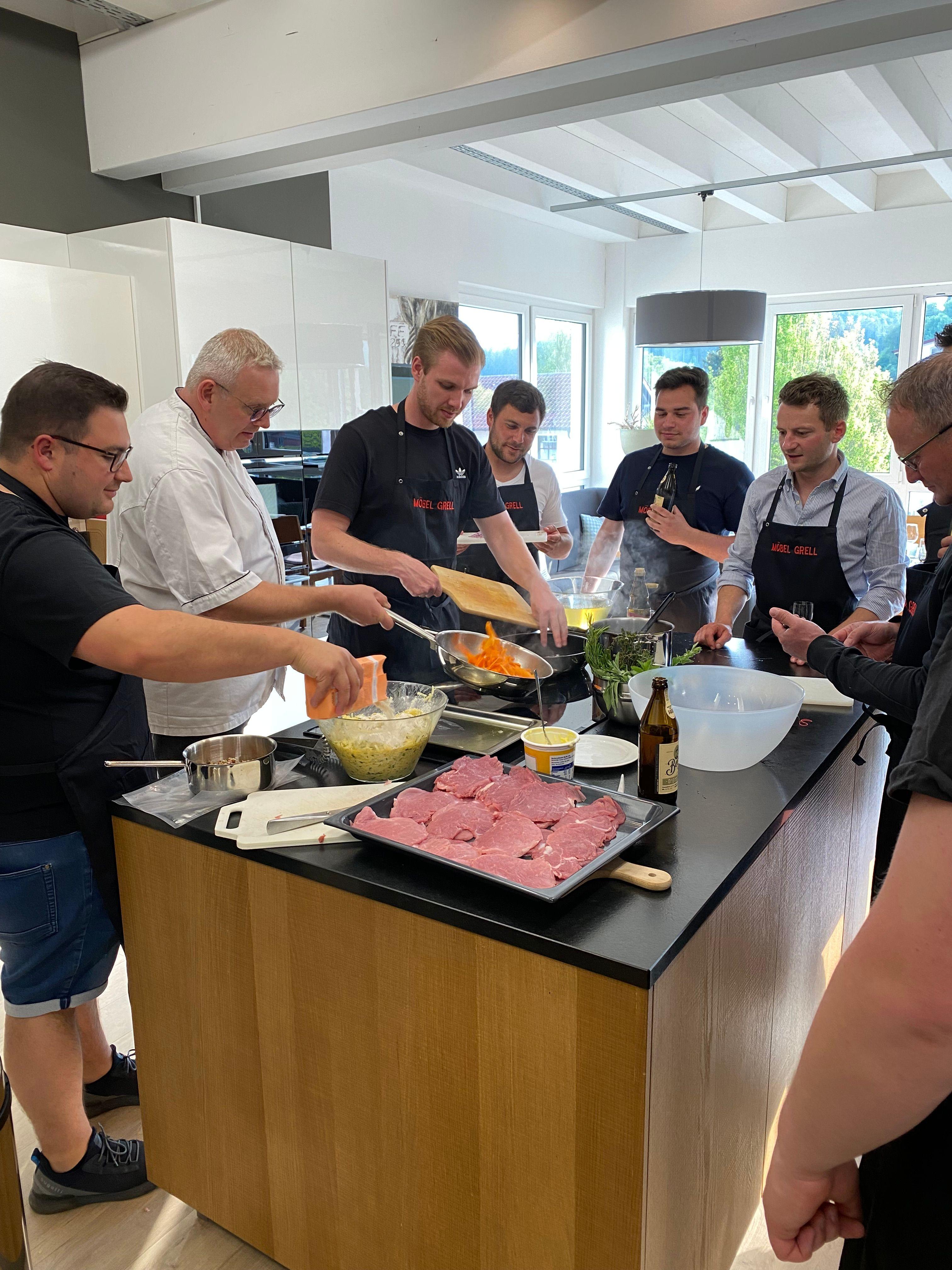 Gruppe von Männern bei Kochkurs in moderner Küche, Koch in schwarzer Schürze bereitet Fleischgericht zu, rohes Fleisch und Zutaten auf Kücheninsel.