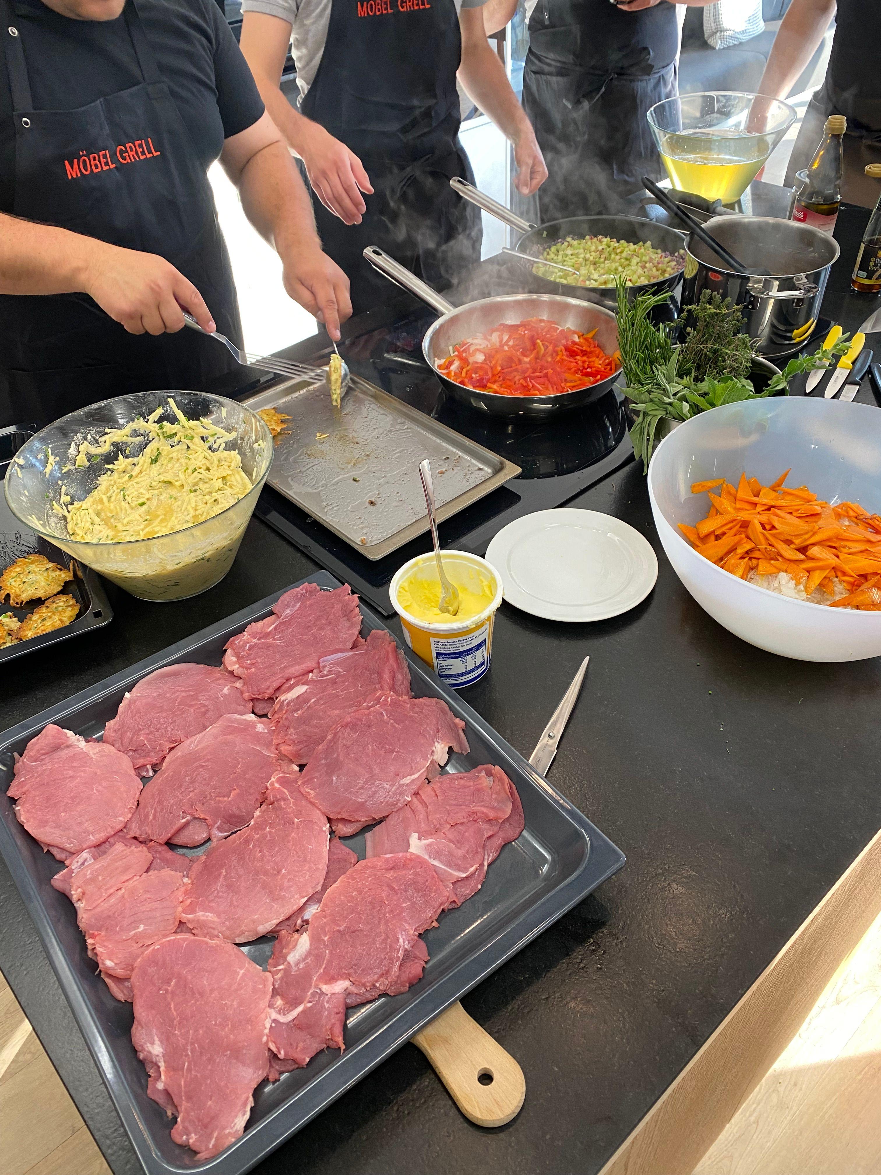 Kochkurs in Aktion: Mehrere Köche in schwarzen "MÖBEL GRELL" Schürzen bereiten gemeinsam Essen zu. Rohes Rindfleisch auf Tablett im Vordergrund, dampfende Pfannen mit Gemüse, Schüsseln mit Zutaten wie Käse und Karotten, frische Kräuter auf dunkler Arbeitsplatte.