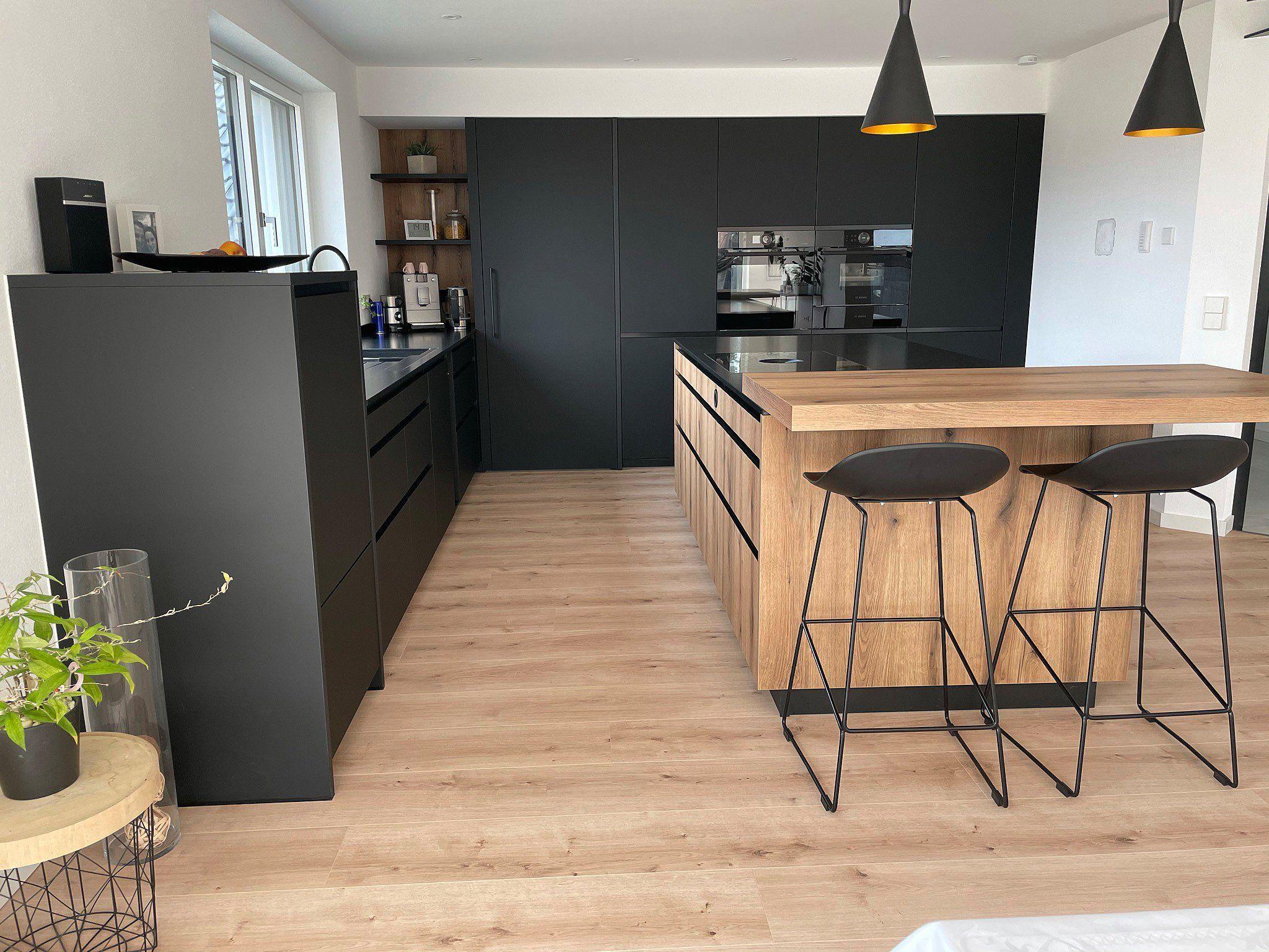 Offene Küche mit dunklen Fronten, Holzkücheninsel, schwarzen Barhockern und rundem Beistelltisch mit Pflanze.