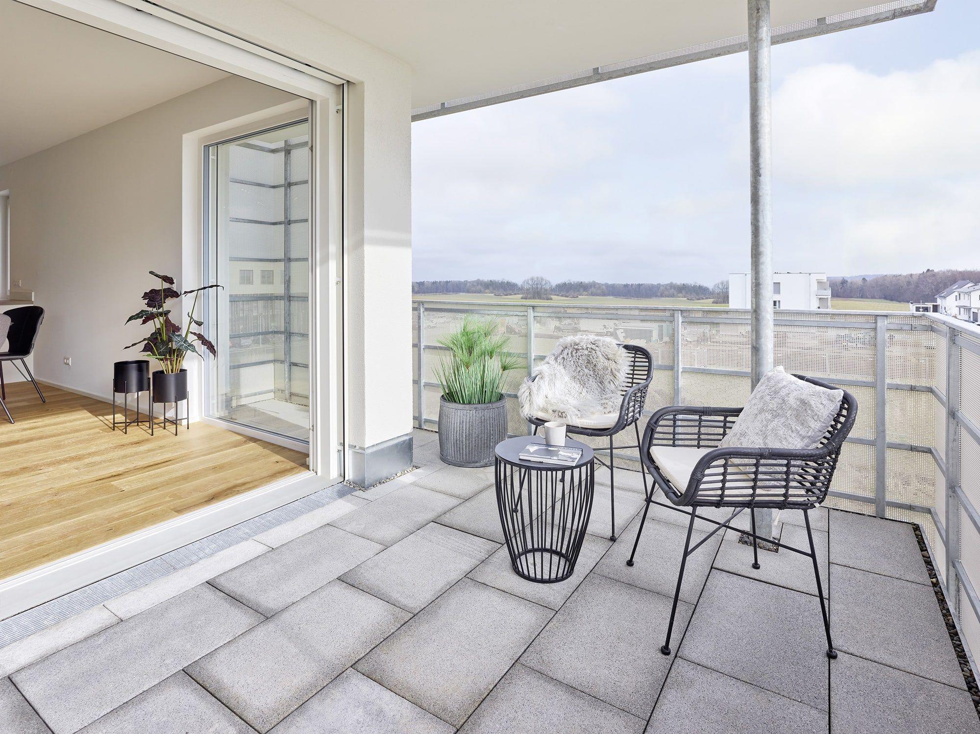 Balkon mit schwarzen Korbstühlen, rundem Beistelltisch, Pflanzen in Töpfen und Glasgeländer, Blick auf Landschaft und moderne Gebäude.