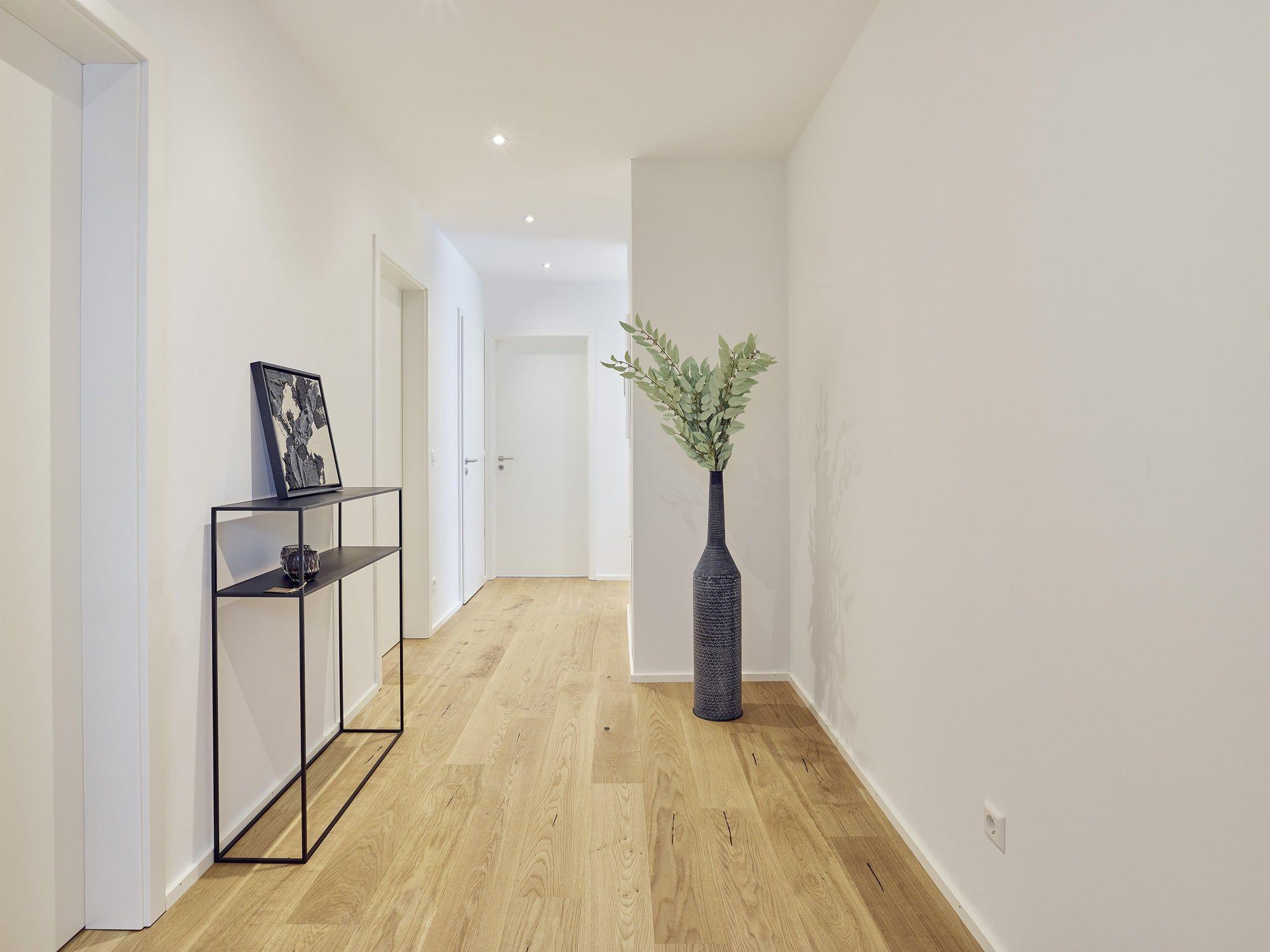 Minimalistischer Flur mit weißen Wänden, Parkett, schwarzem Metallregal mit Dekoration und großer schwarzer Bodenvase mit grünen Zweigen.