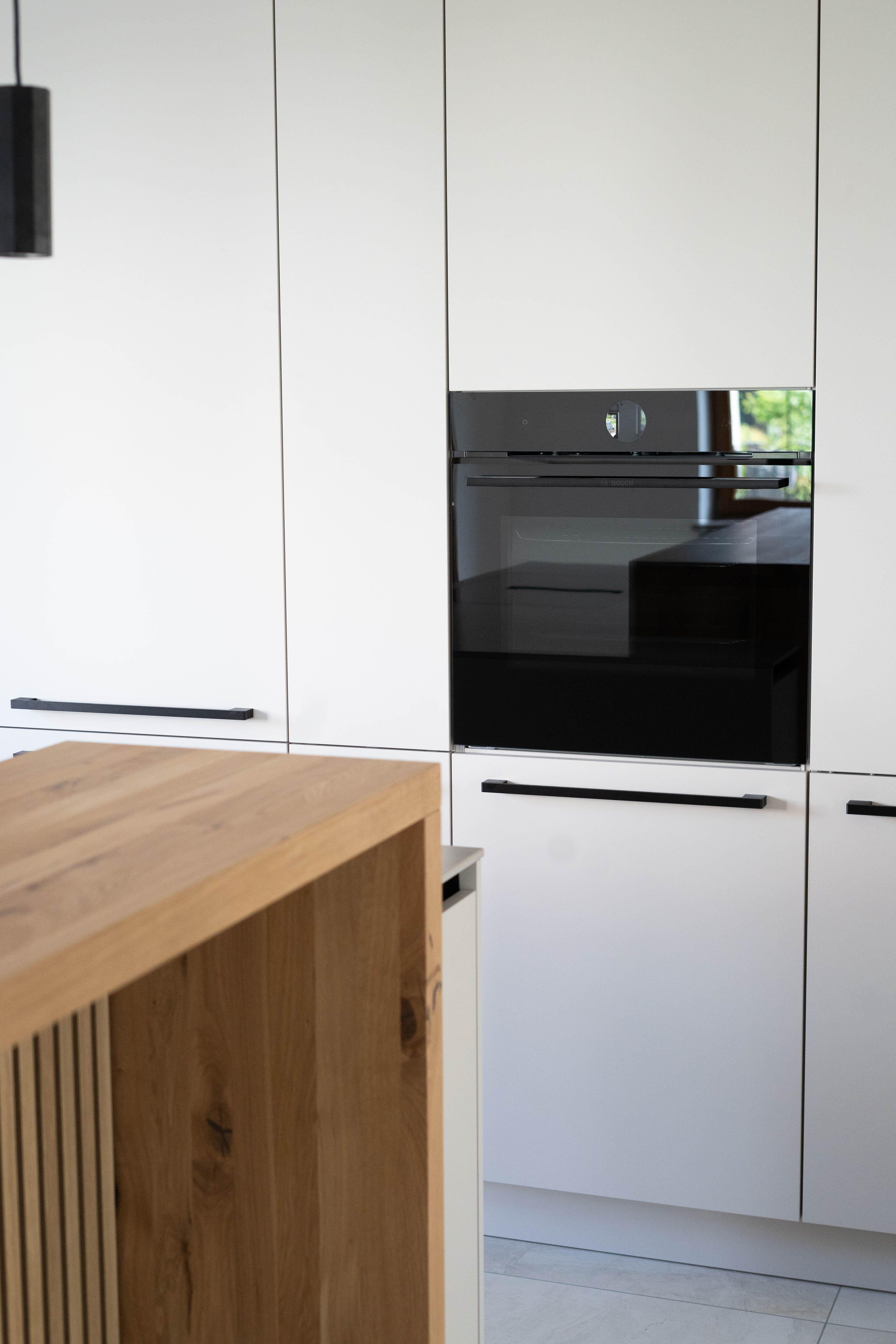 Weißer Hochschrank mit eingebautem Backofen neben Holztheke und Lamellenfront der Kücheninsel.