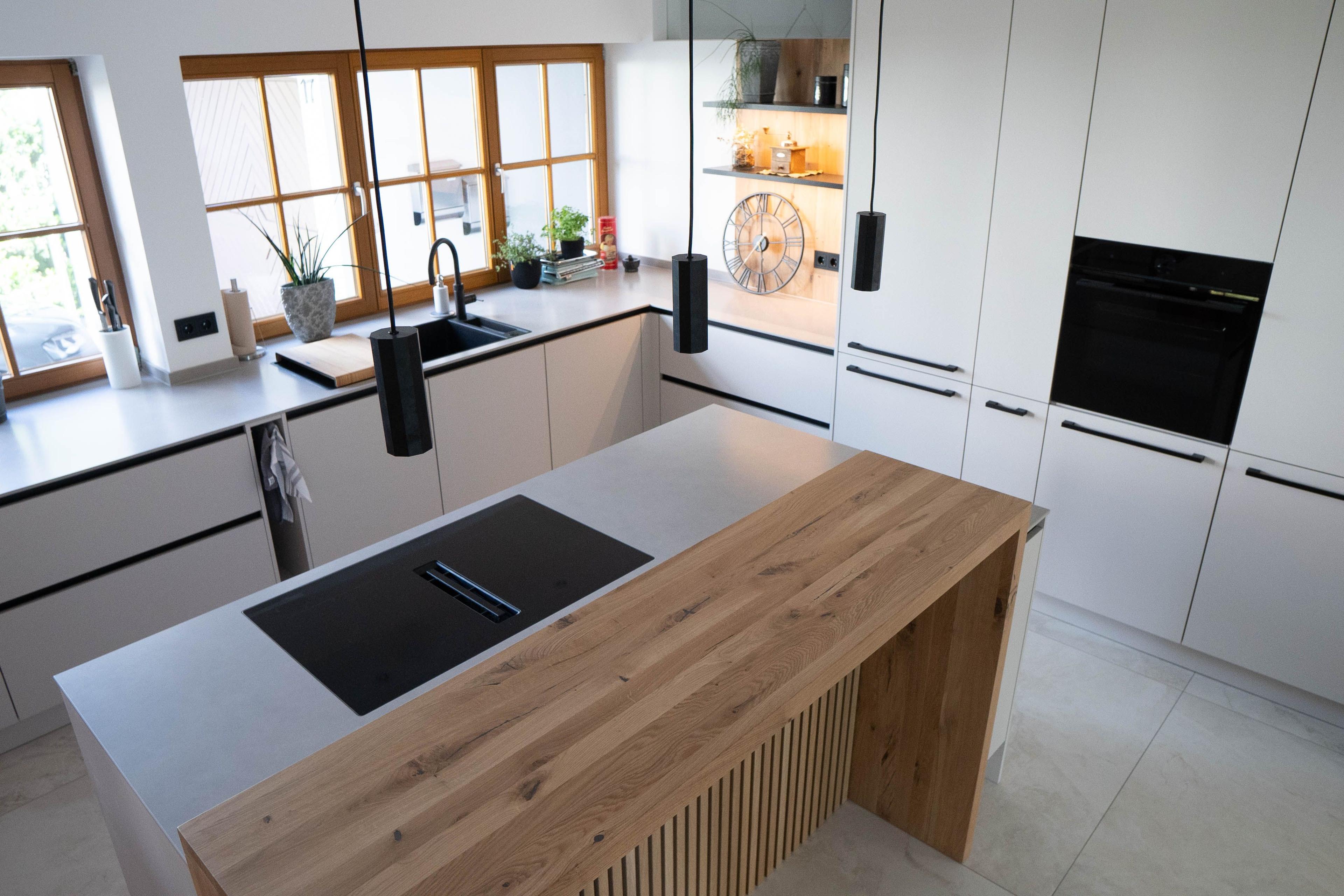 Moderne weiße Küche mit heller Insel, Holztheke, Lamellenfront, schwarzem Kochfeld und offenem Regal mit Beleuchtung.