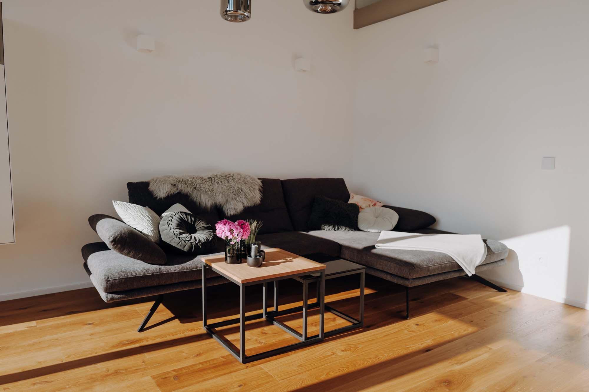 Wohnbereich mit grauer Eckcouch, Metallcouchtisch, bunten Kissen und modernen Pendelleuchten über dem Esstisch.