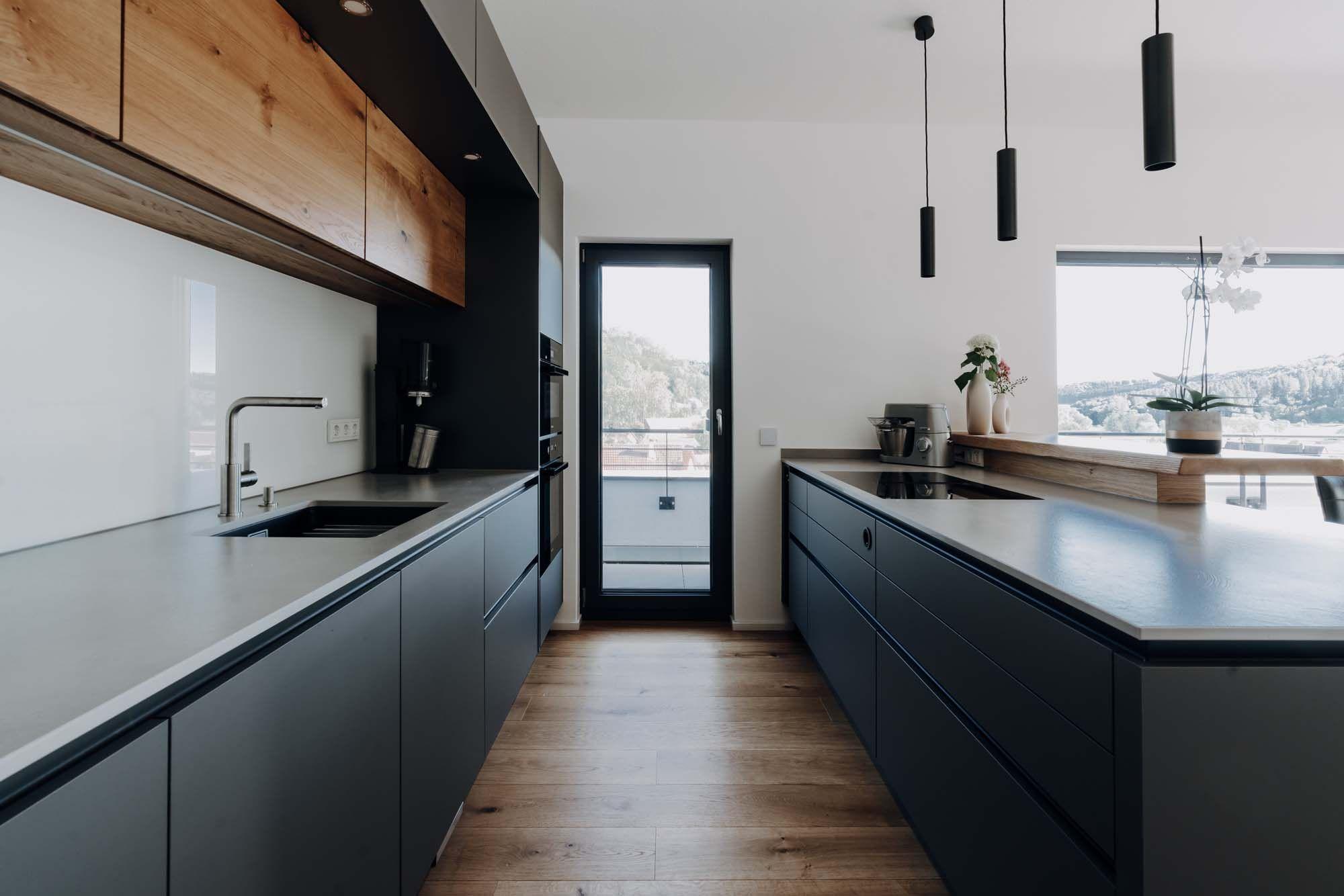 Moderne Küche in Grautönen mit Holzoberschränken, weißer Arbeitsplatte, schwarzen Pendelleuchten und Zugang zum Balkon.