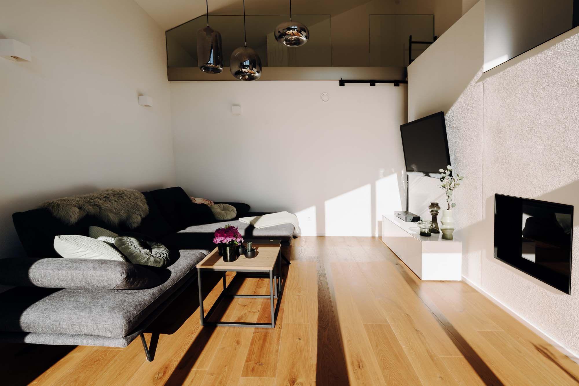 Wohnbereich mit grauem Ecksofa, Fellkissen, schwarzen Pendelleuchten und Holzboden.