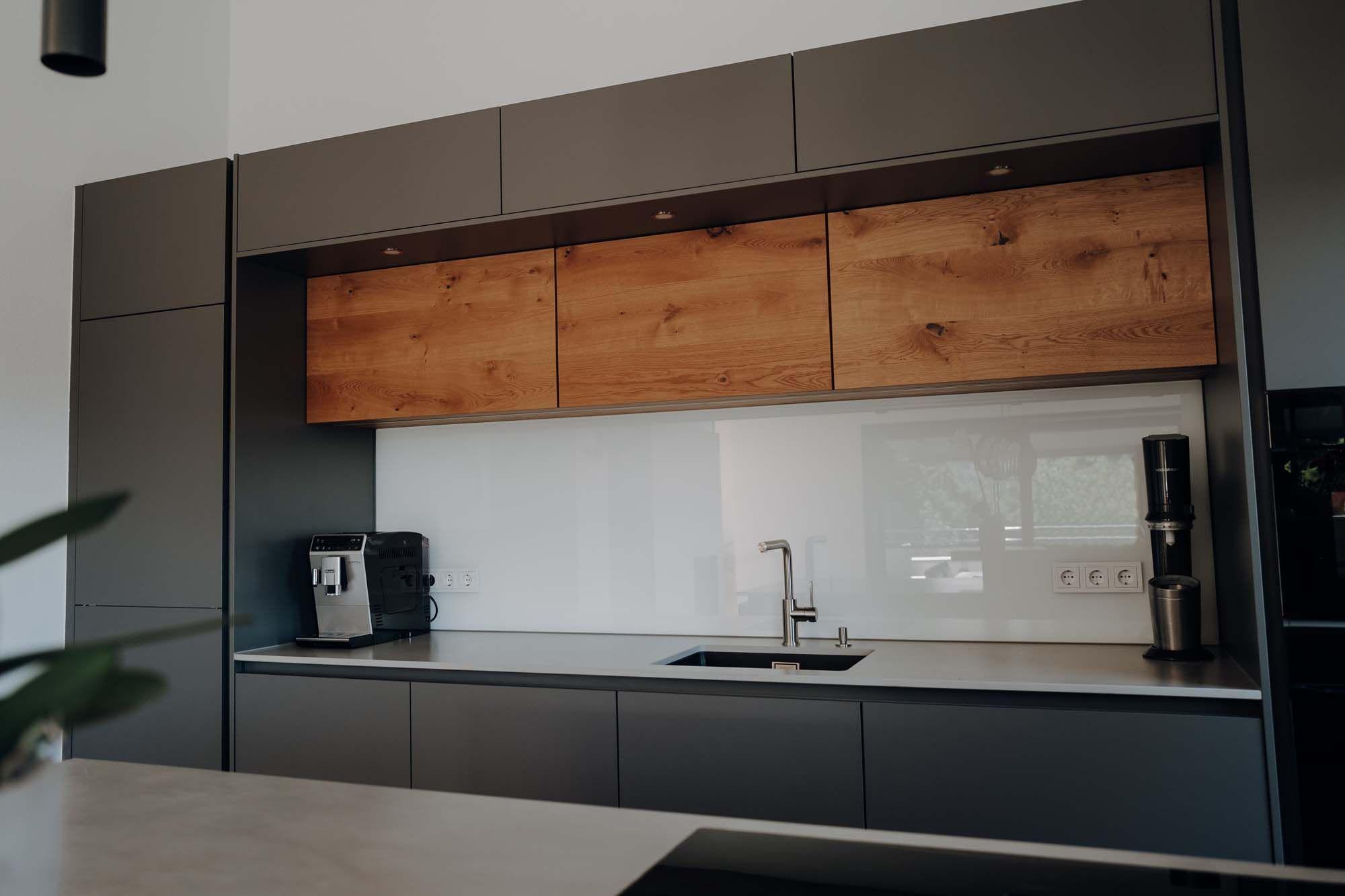 Küchenwand mit anthrazitfarbenen Unterschränken, Holz-Hängeschränken, weißer Glasrückwand und Kaffeemaschine.