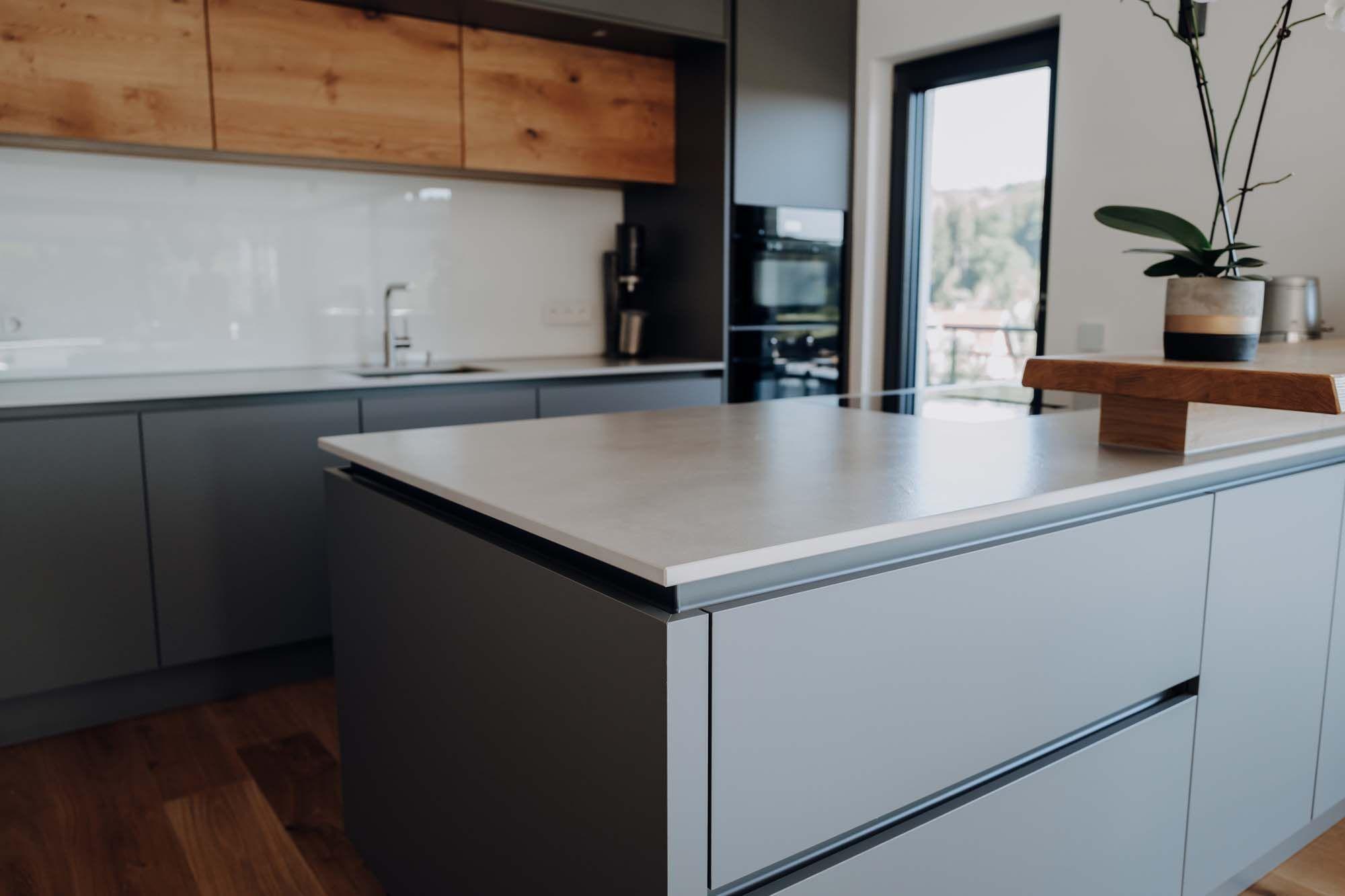 ücheninsel-Detailansicht mit weißer Arbeitsplatte, grauen Fronten, Holzelement und Orchideen-Dekoration im Hintergrund.