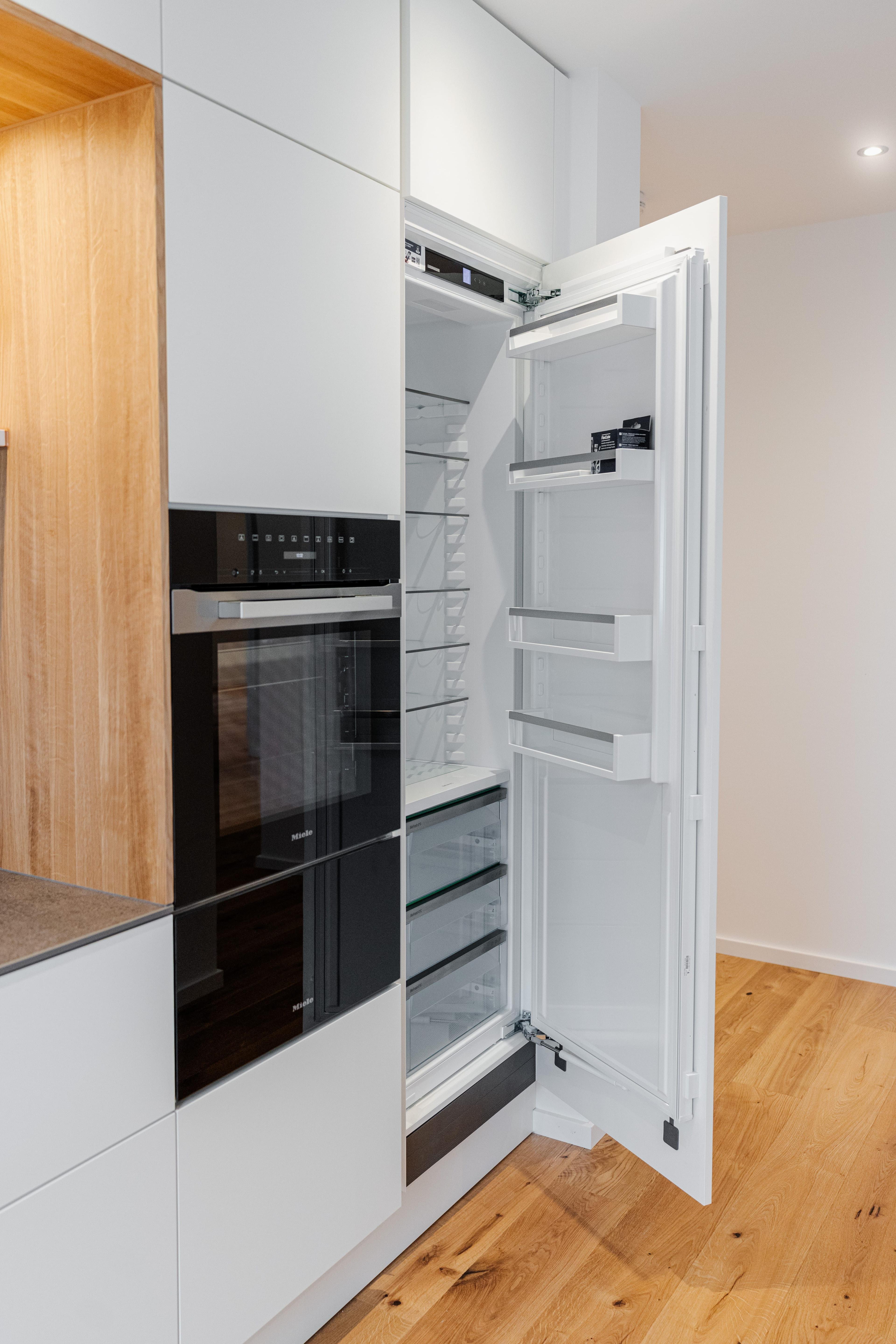 Geöffneter Einbaukühlschrank in weißer Küche mit Holznische, eingebauten Geräten und Eichenparkett.
