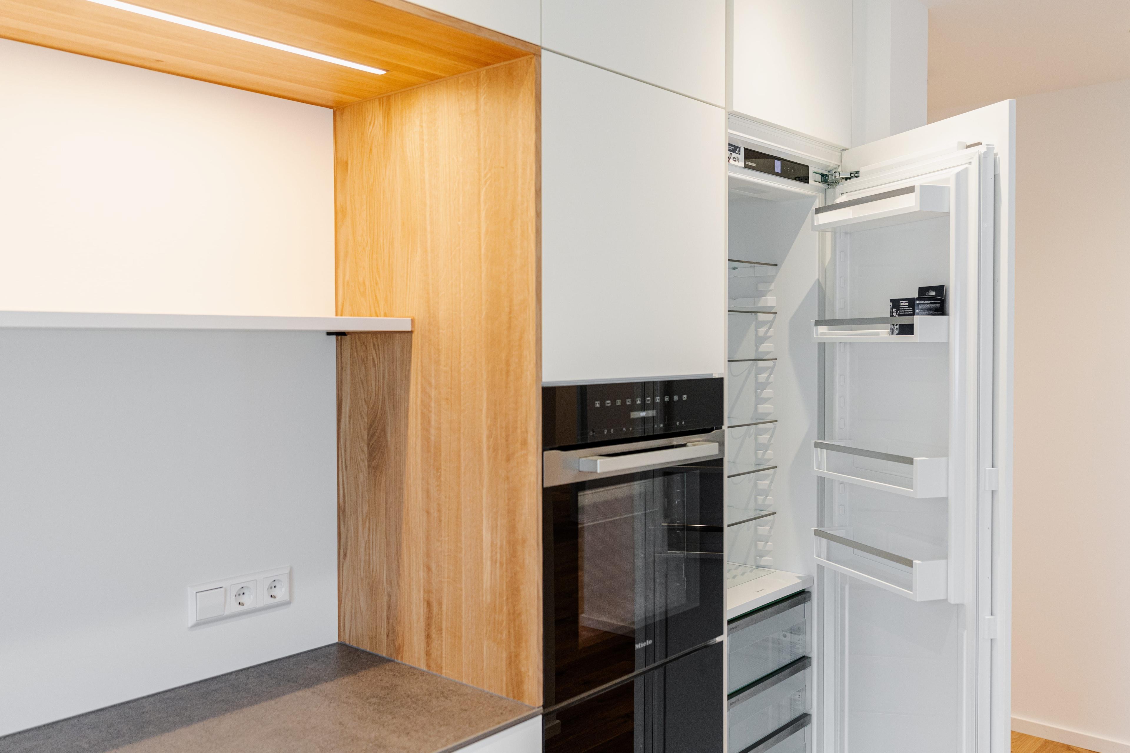 Kühlschrank mit geöffneter Tür neben beleuchteter Holznische, weißen Küchenfronten und grauer Arbeitsplatte.