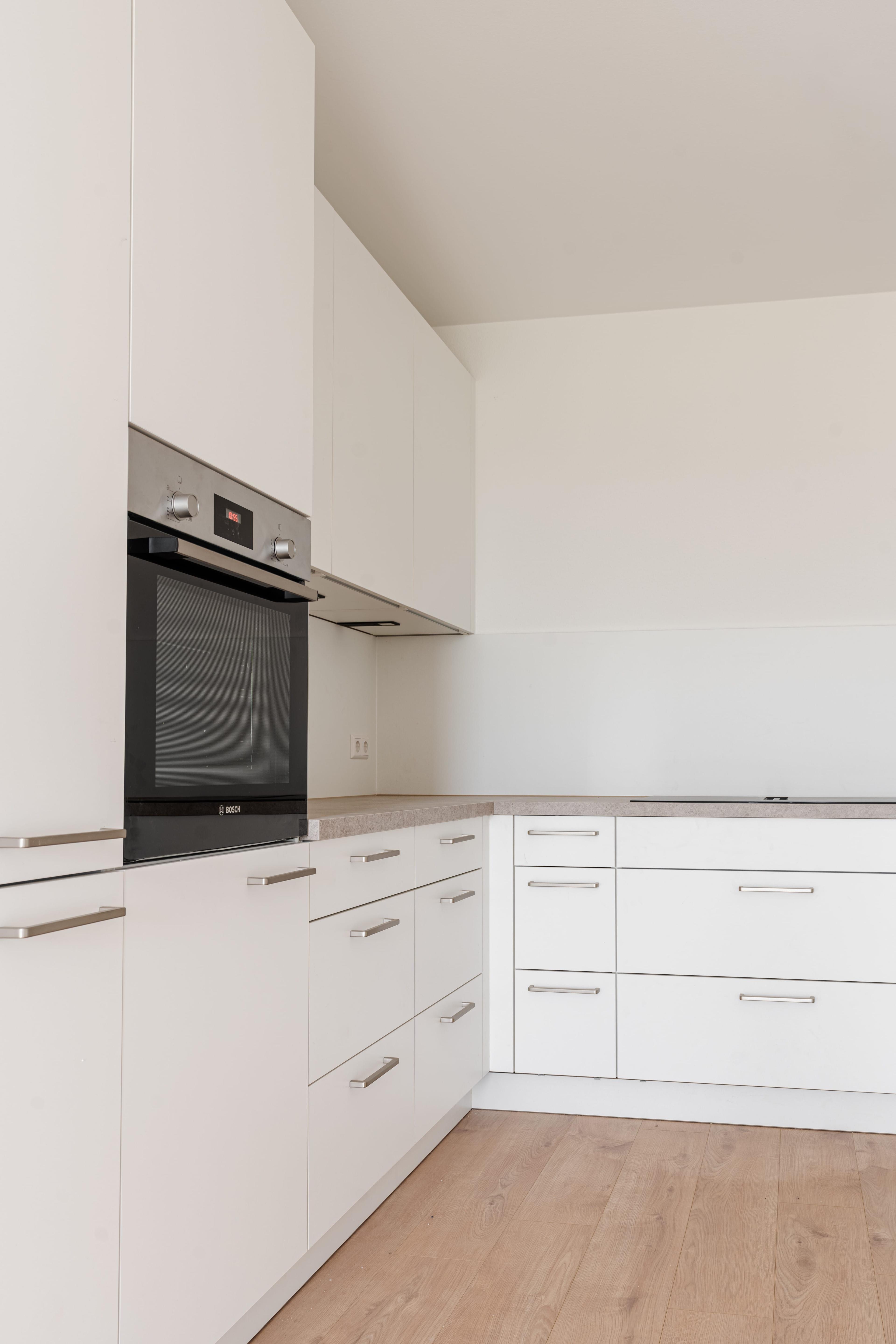Küchenzeile mit weißen grifflosen Fronten, eingebautem Backofen, beiger Steinarbeitsplatte und Holzparkett.