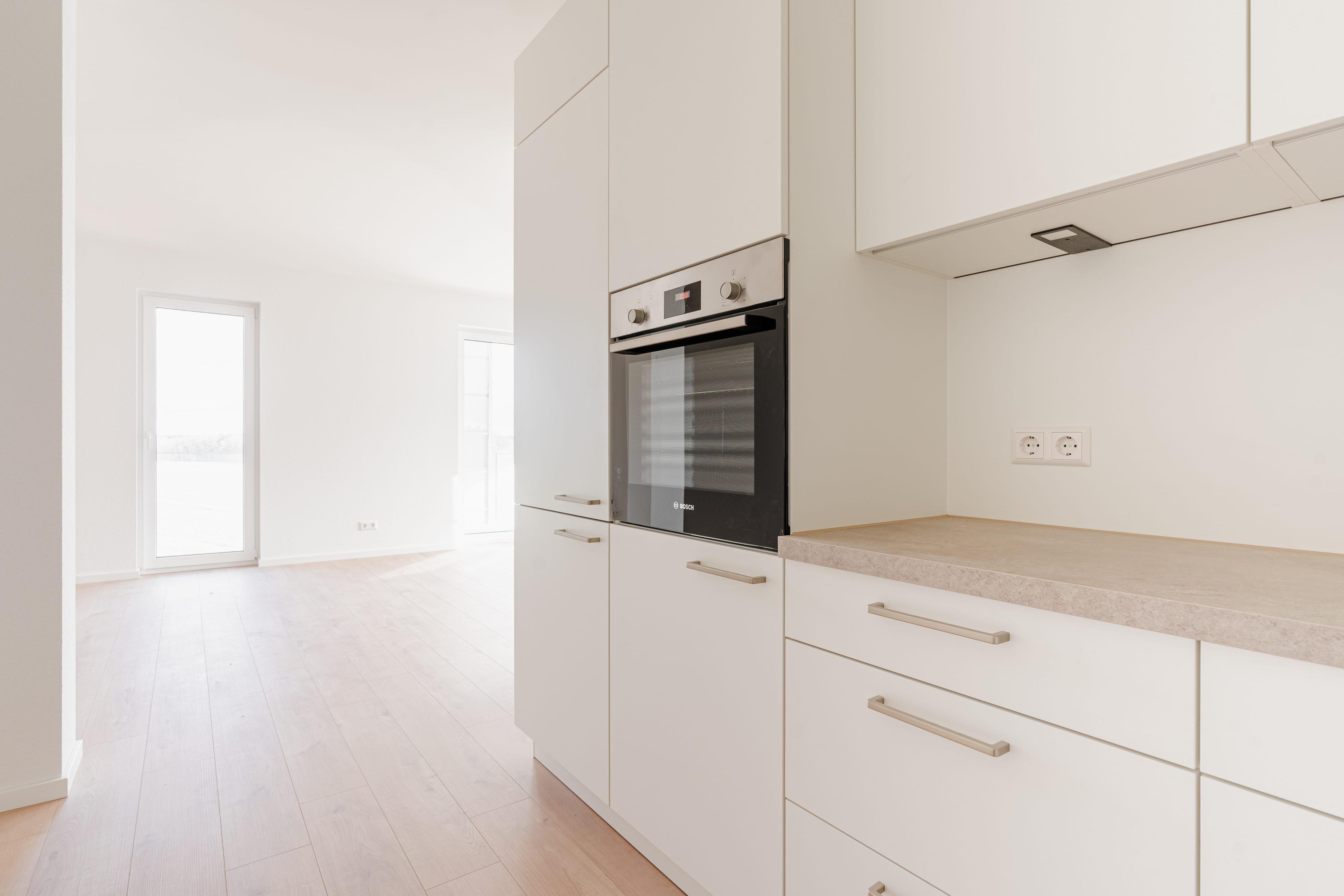 Küchenzeile mit eingebautem Backofen, weißen Fronten, beiger Arbeitsplatte und Blick in angrenzenden Raum.