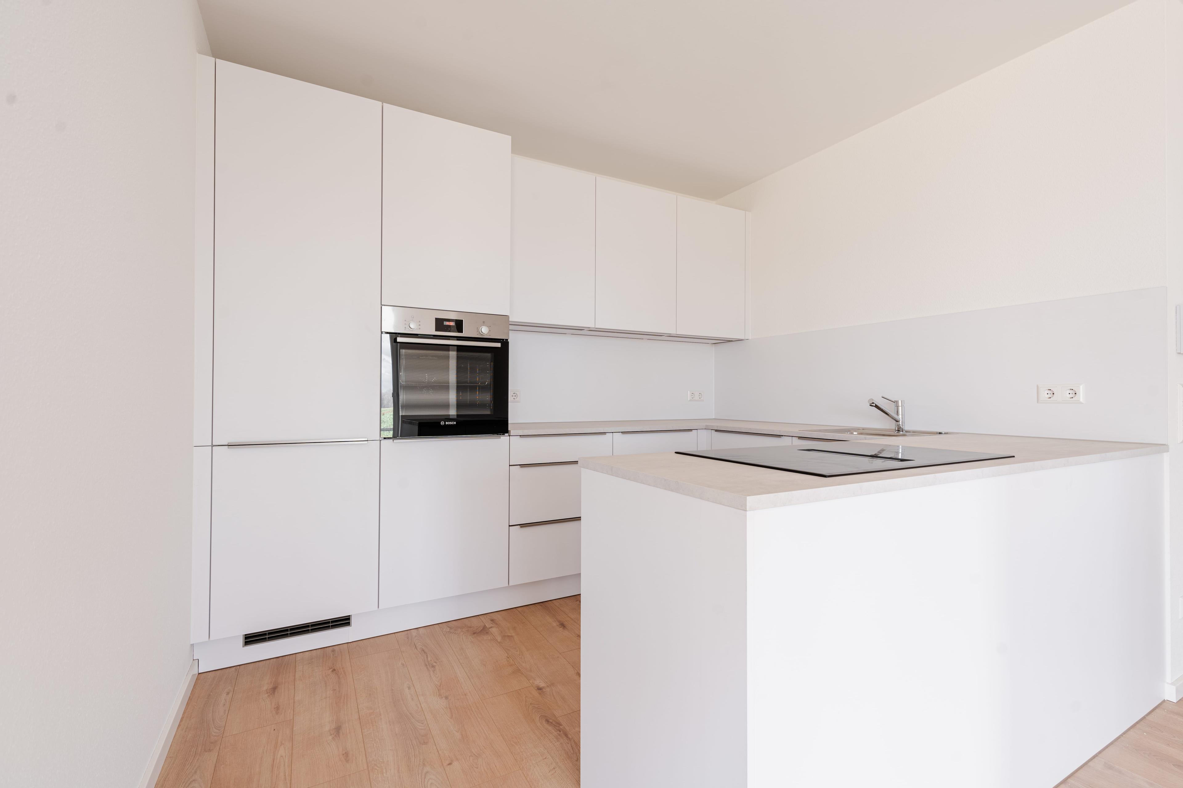Moderne weiße Küche mit Kochinsel, eingebautem Backofen und durchgehender heller Arbeitsplatte.
